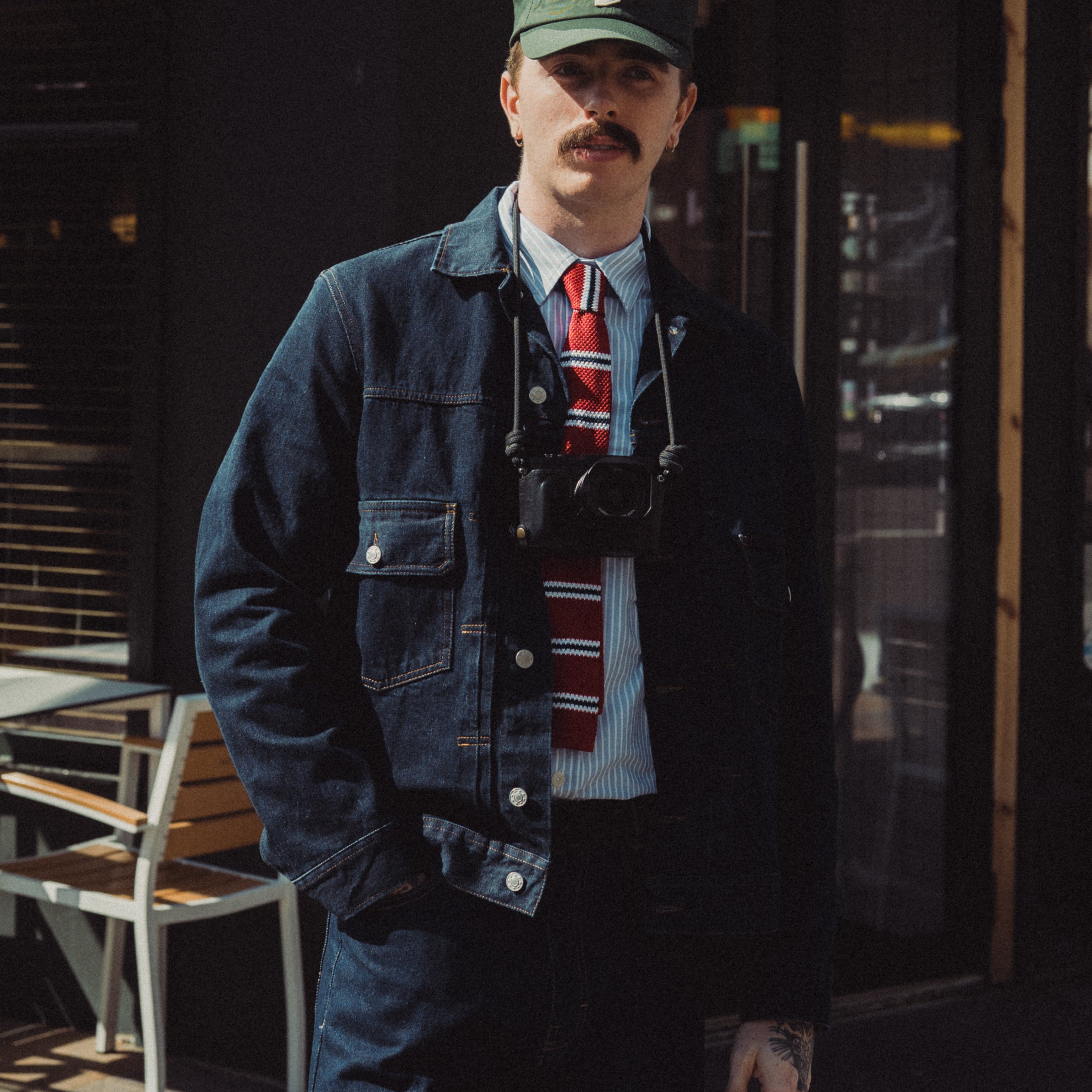 Man wearing a denim jacket, cap, and tie standing outdoors.