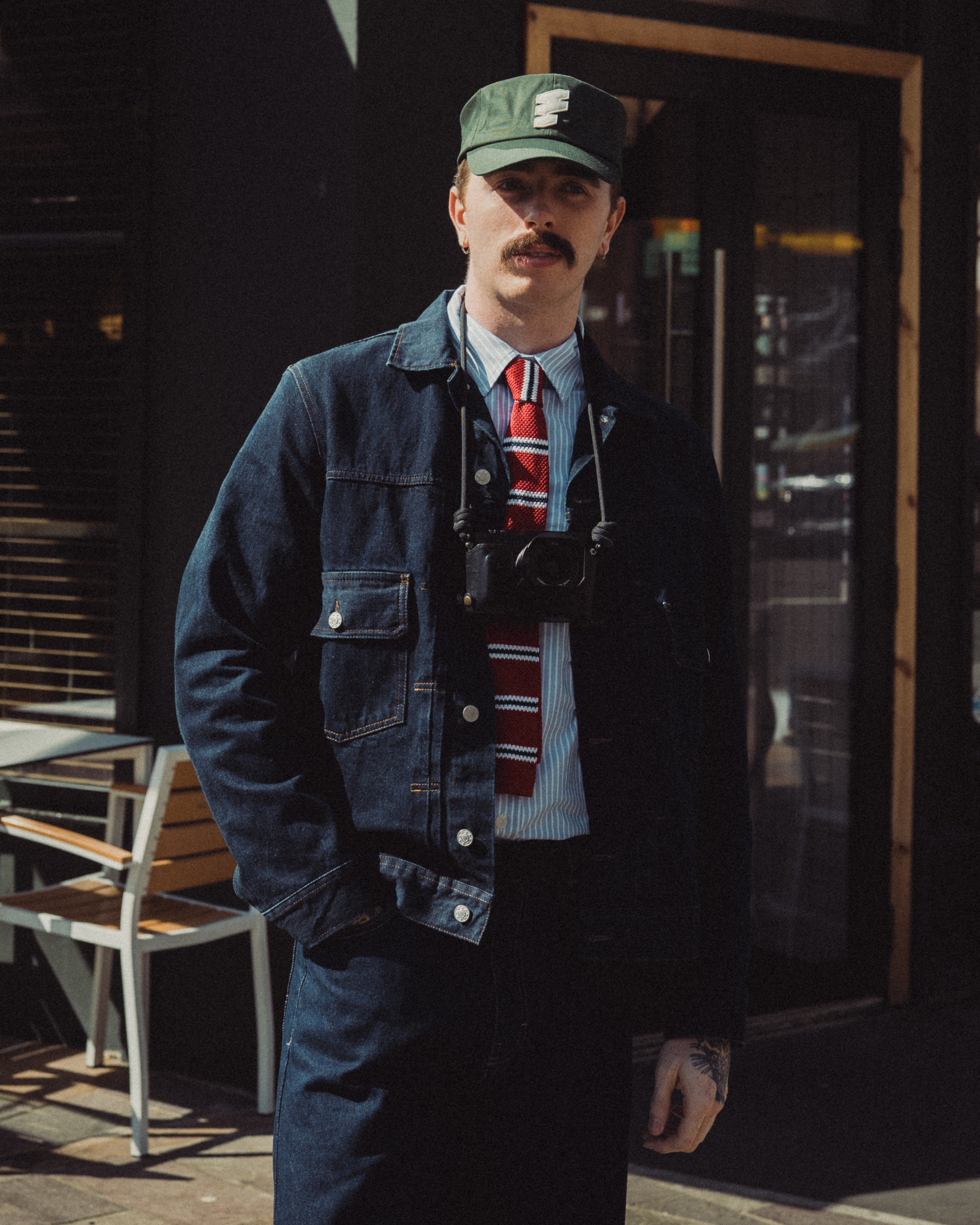 Man wearing a denim jacket, cap, and tie standing outdoors.