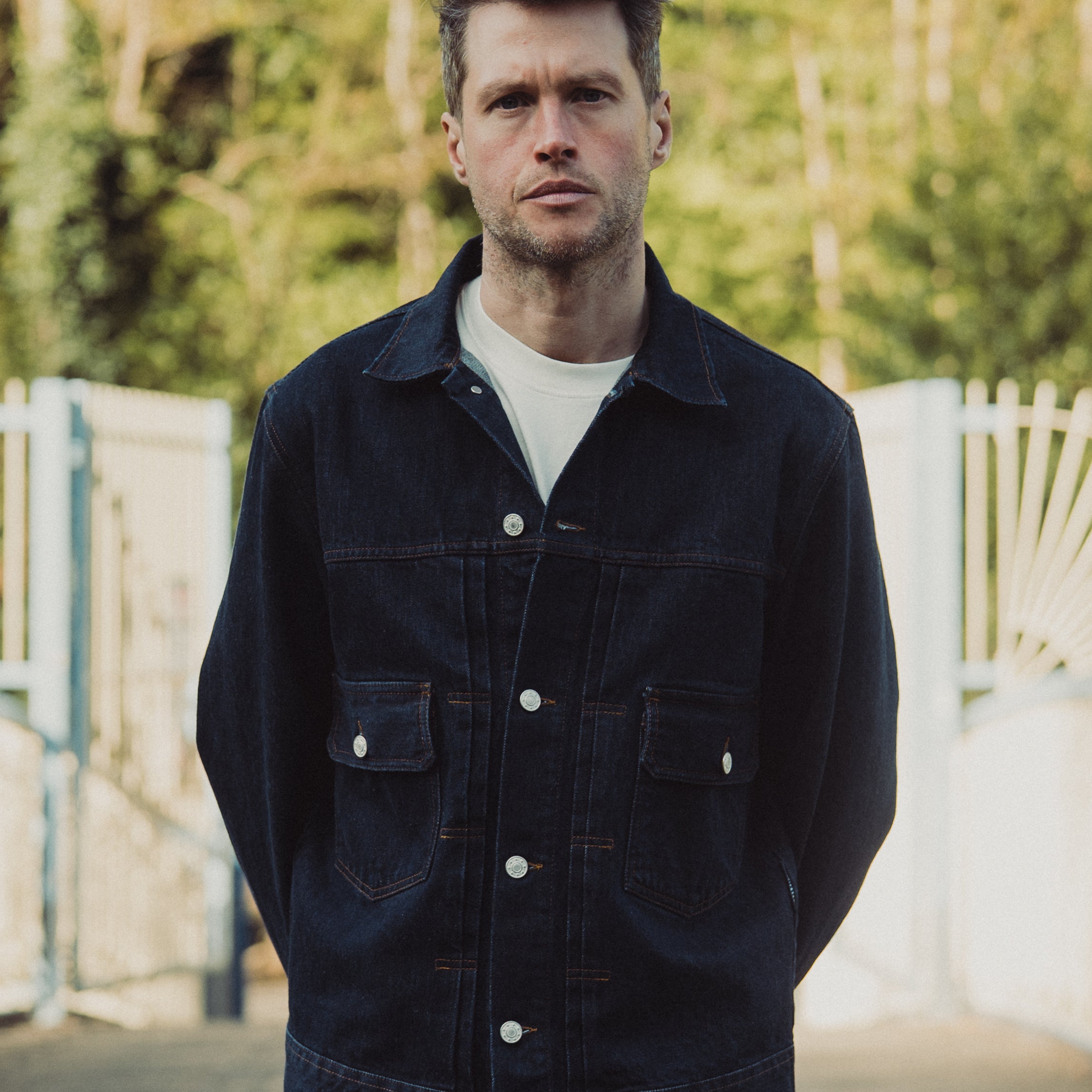 Man wearing a dark denim jacket outdoors with blurred greenery in the background