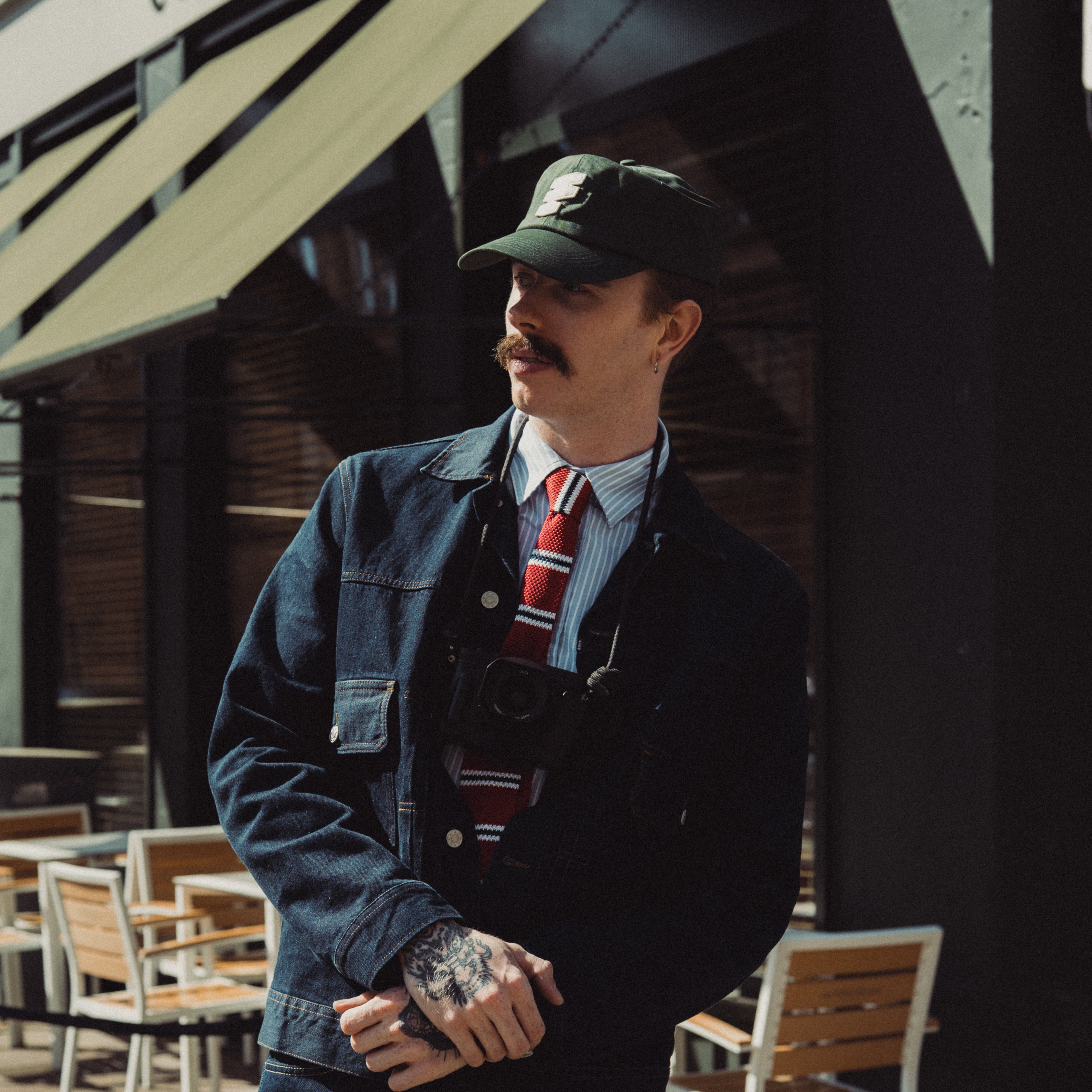 Man in a denim jacket and cap standing in front of a building with an awning.