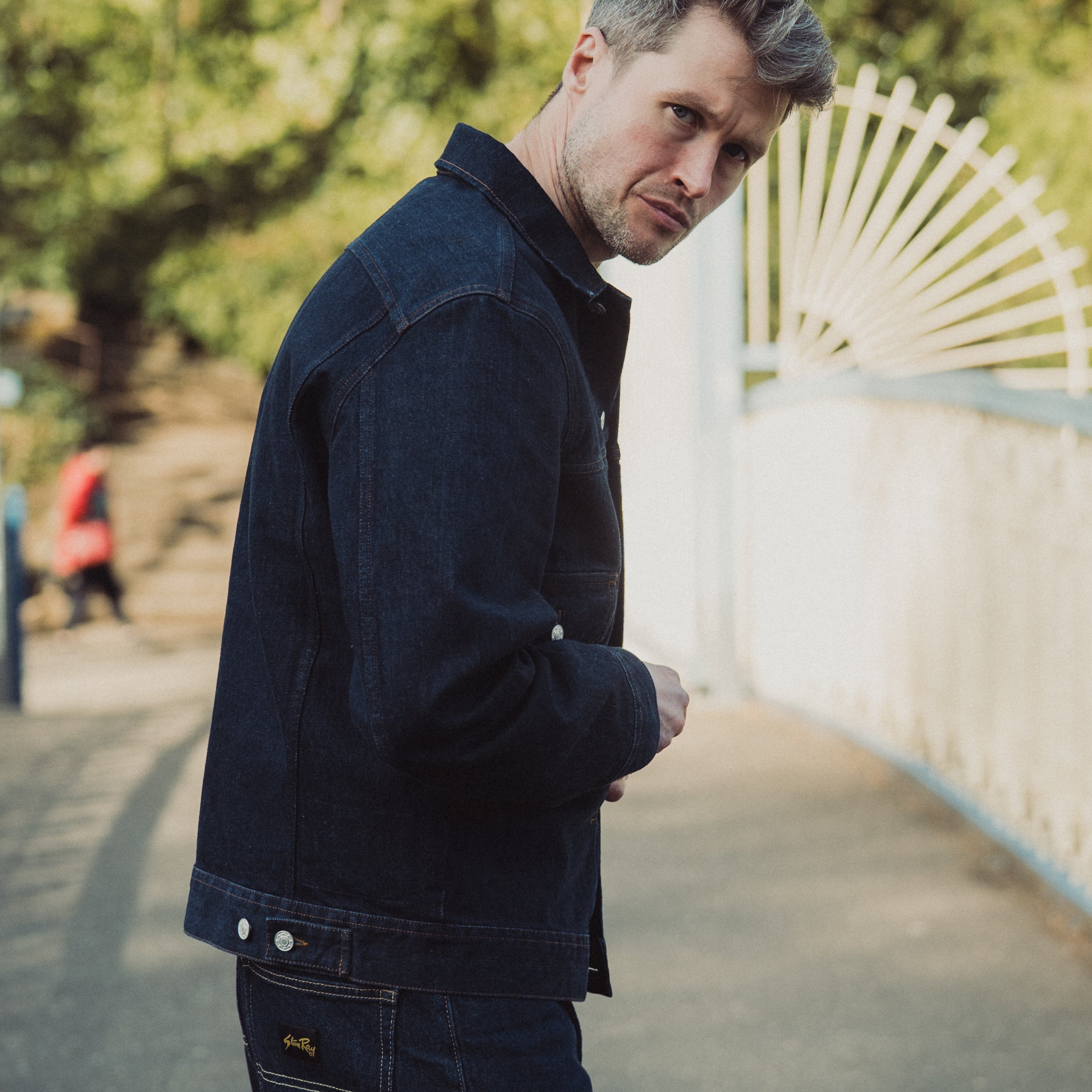 Man wearing a dark jacket and pants standing outdoors with trees and a white structure in the background.