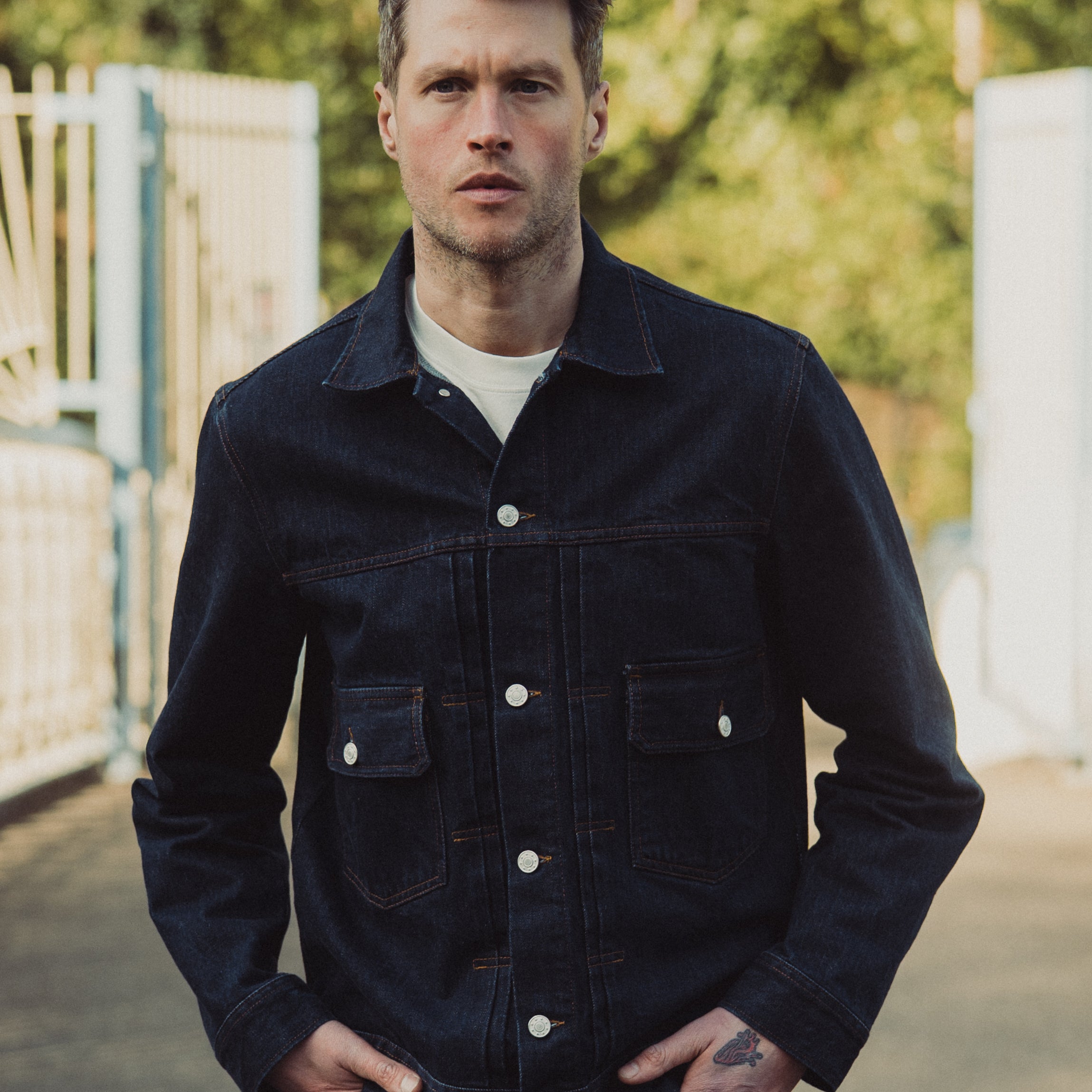 Man wearing a dark denim jacket standing outdoors with greenery in the background
