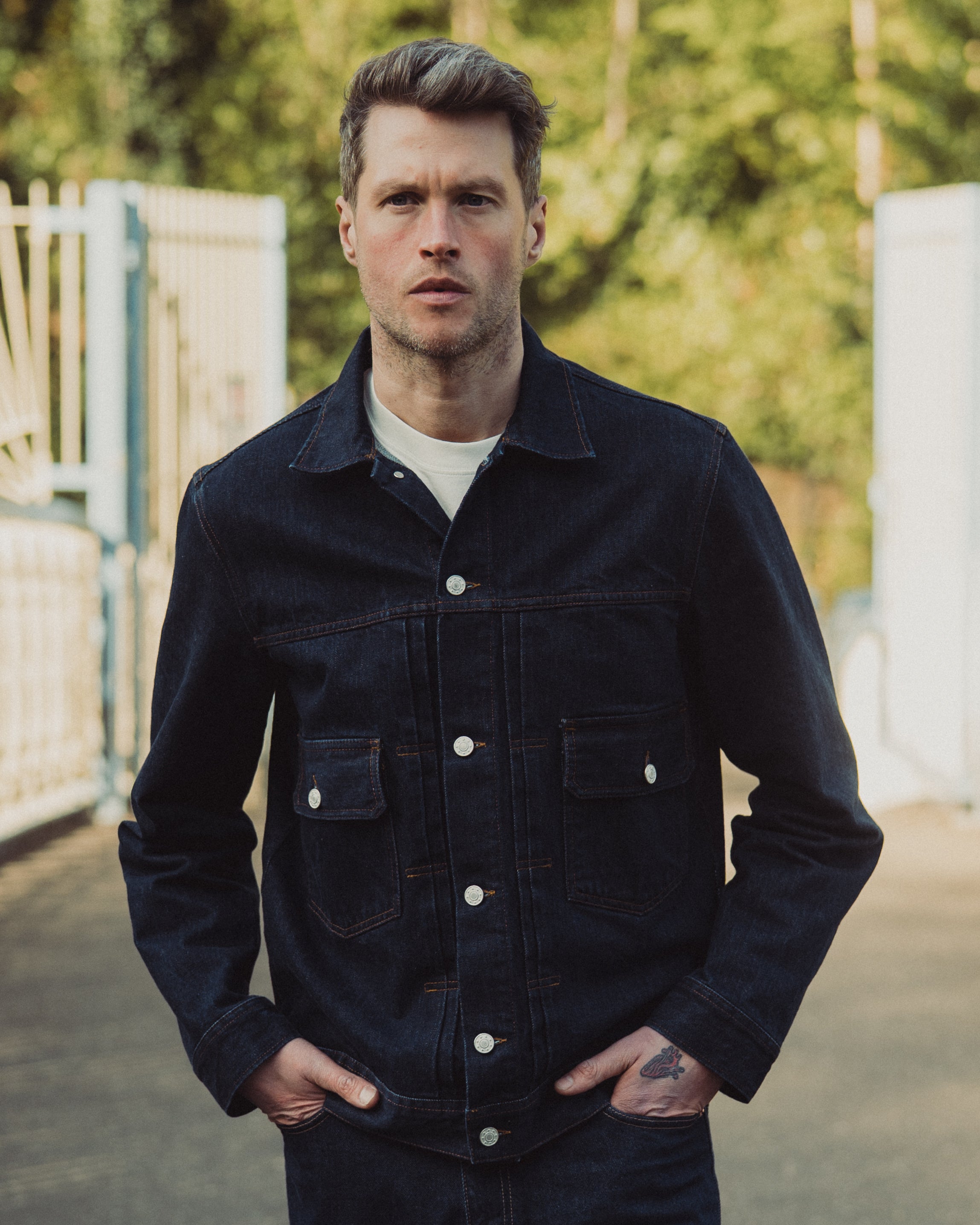 Man wearing a dark denim jacket standing outdoors with greenery in the background