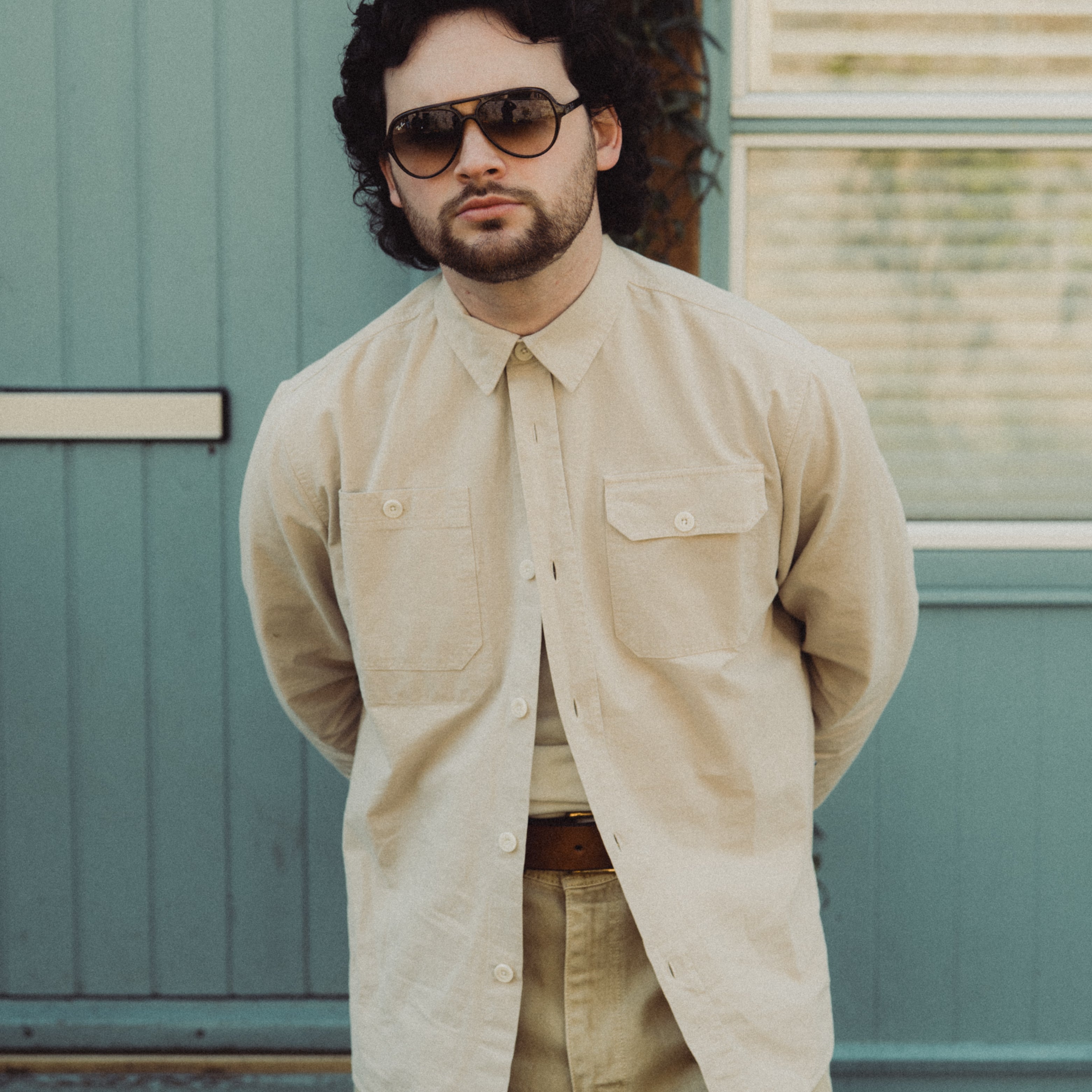 Man wearing sunglasses and a beige shirt standing in front of a teal door.