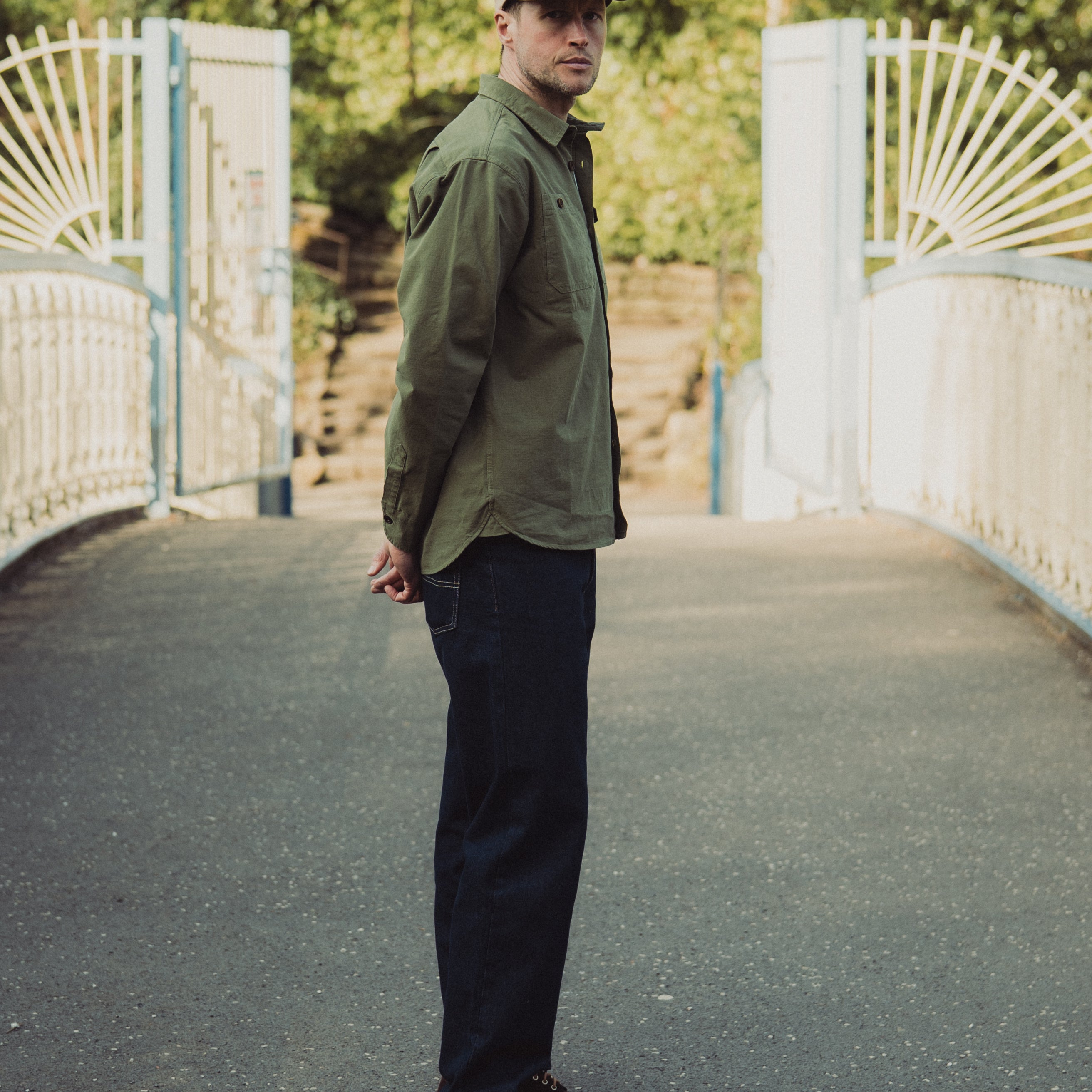 Man wearing a green jacket and cap standing on a bridge with a white gate and trees in the background.