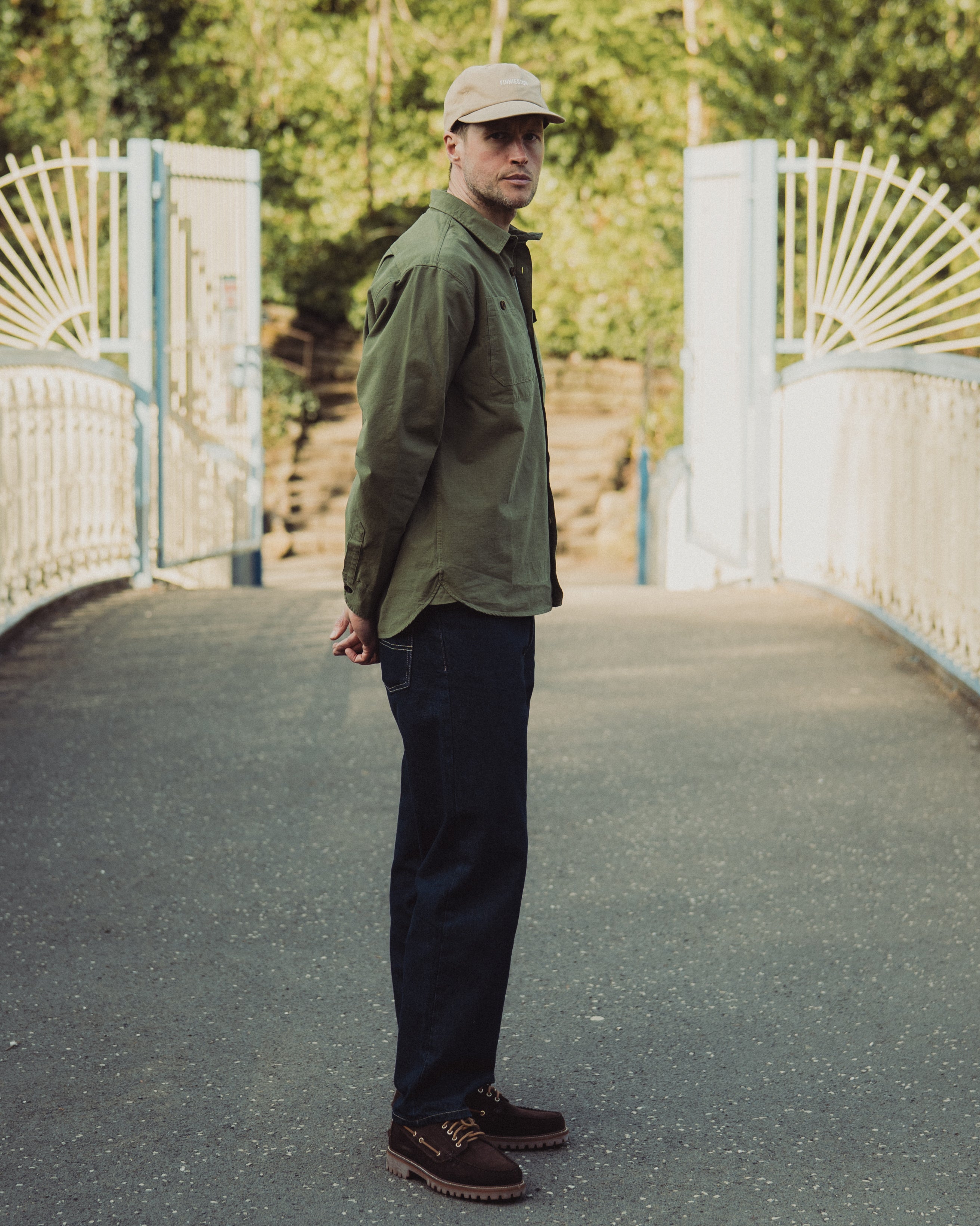 Man wearing a green jacket and cap standing on a bridge with a white gate and trees in the background.