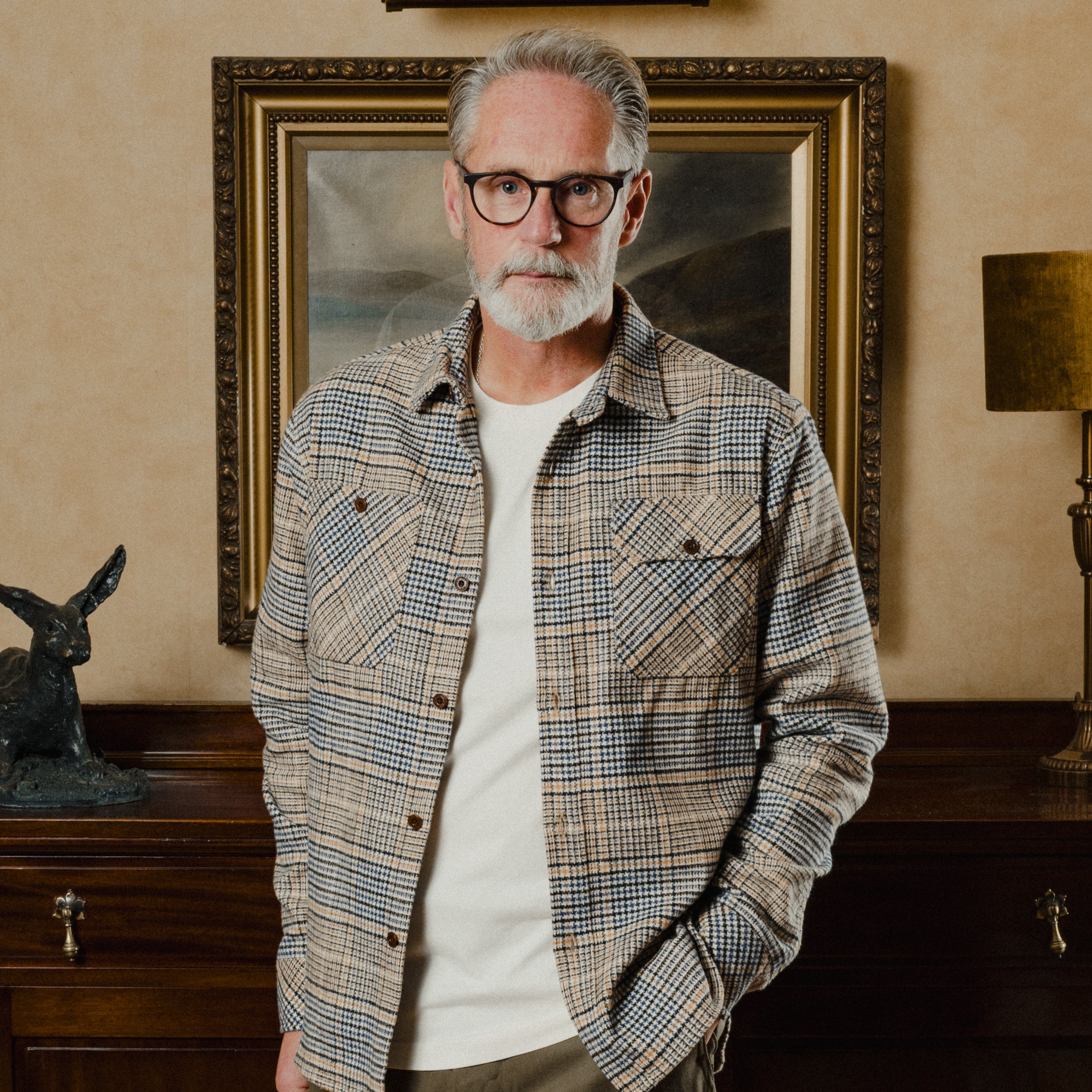 Man wearing a plaid shirt and glasses standing in a room with a painting and lamp.