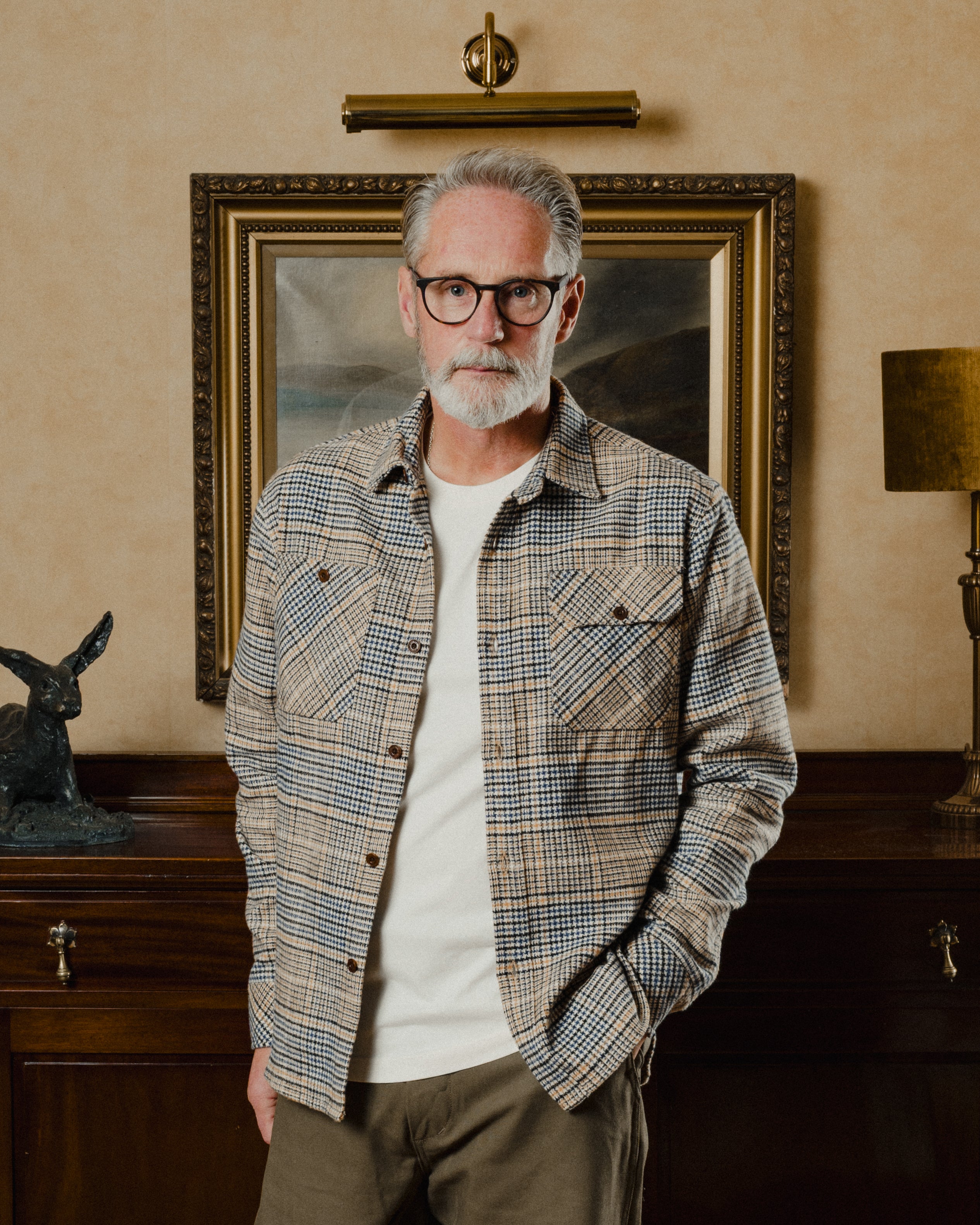 Man wearing a plaid shirt and glasses standing in a room with a painting and lamp.