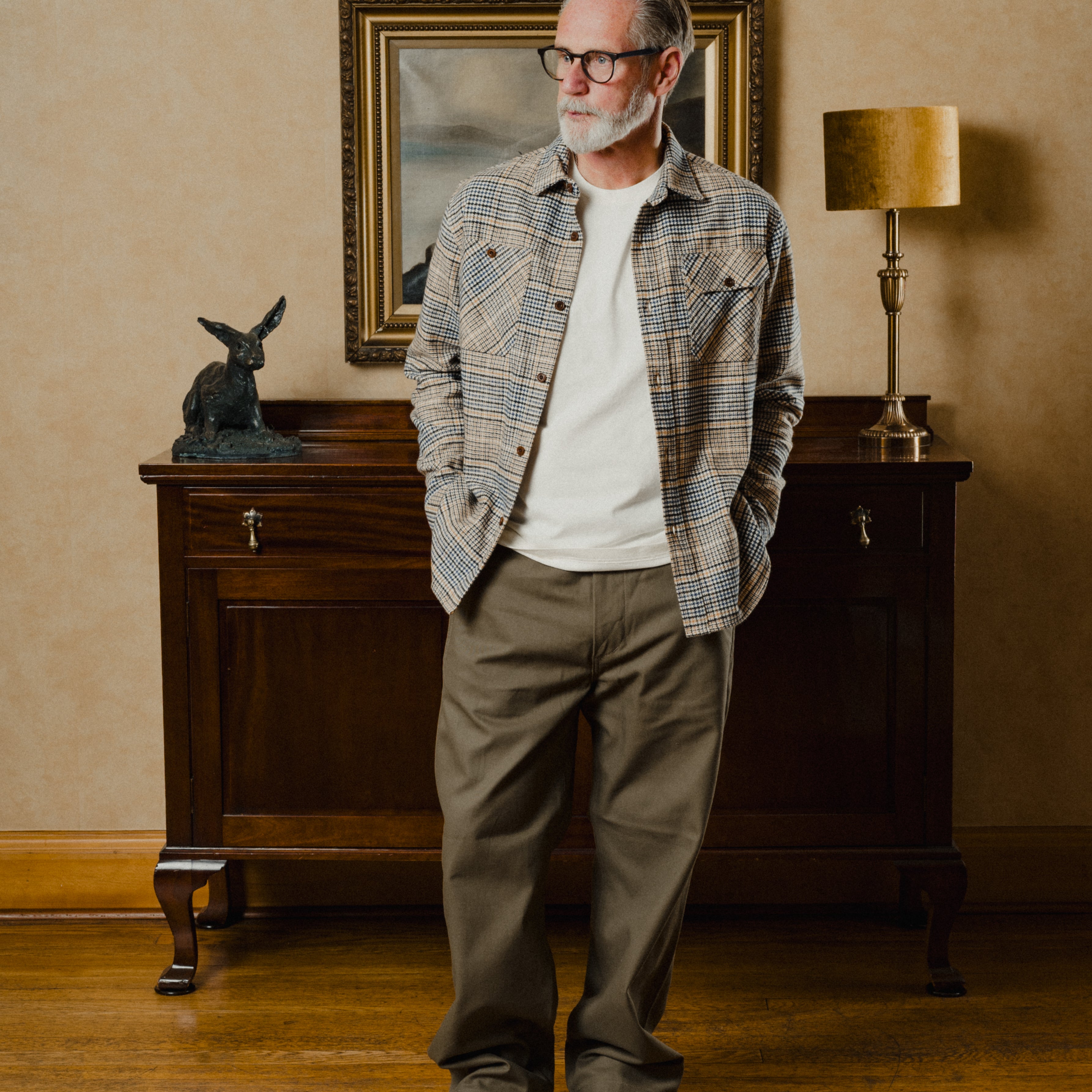 Man standing in a room with a wooden cabinet, lamp, and painting in the background.