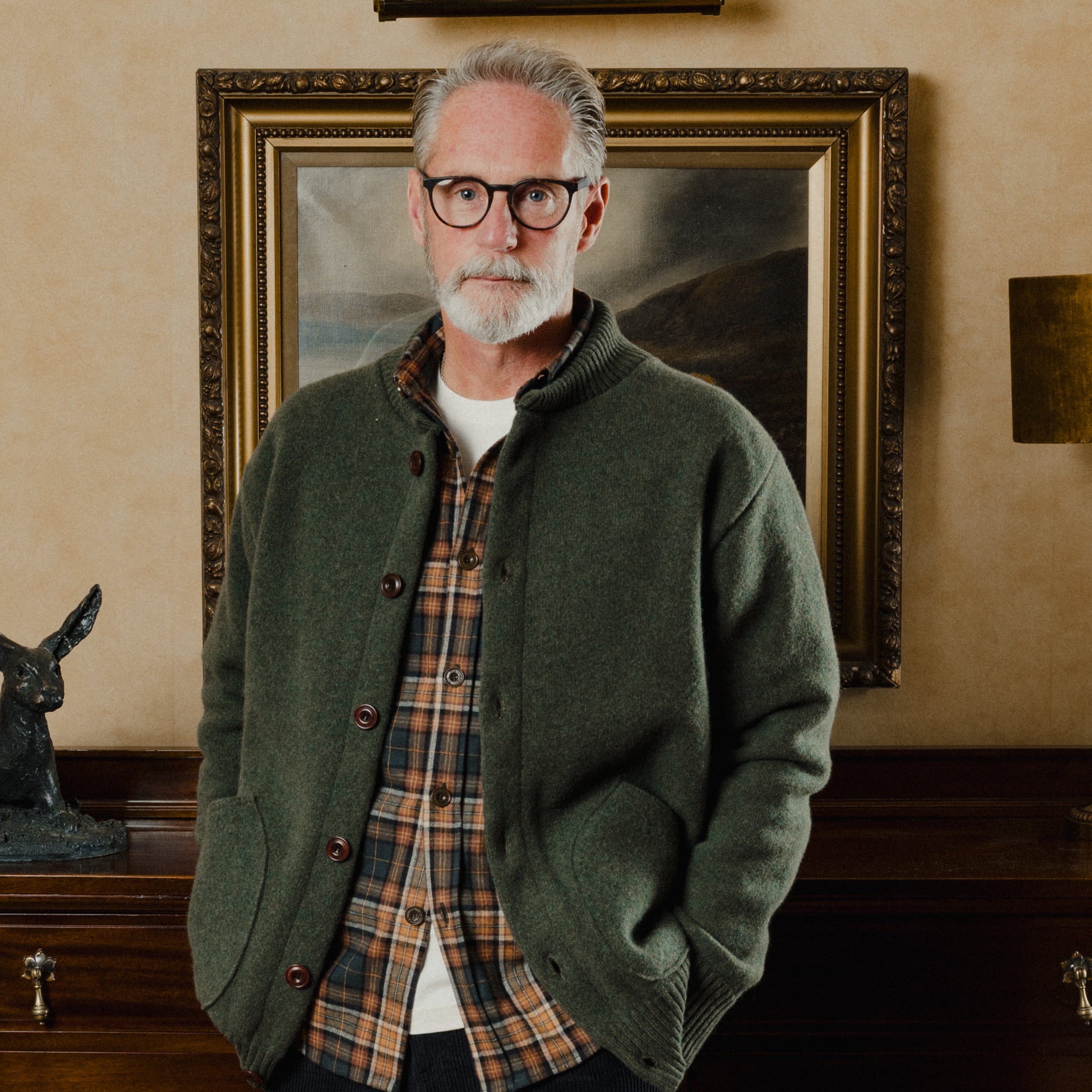 Man wearing a green cardigan over a plaid shirt in a room with a painting on the wall.
