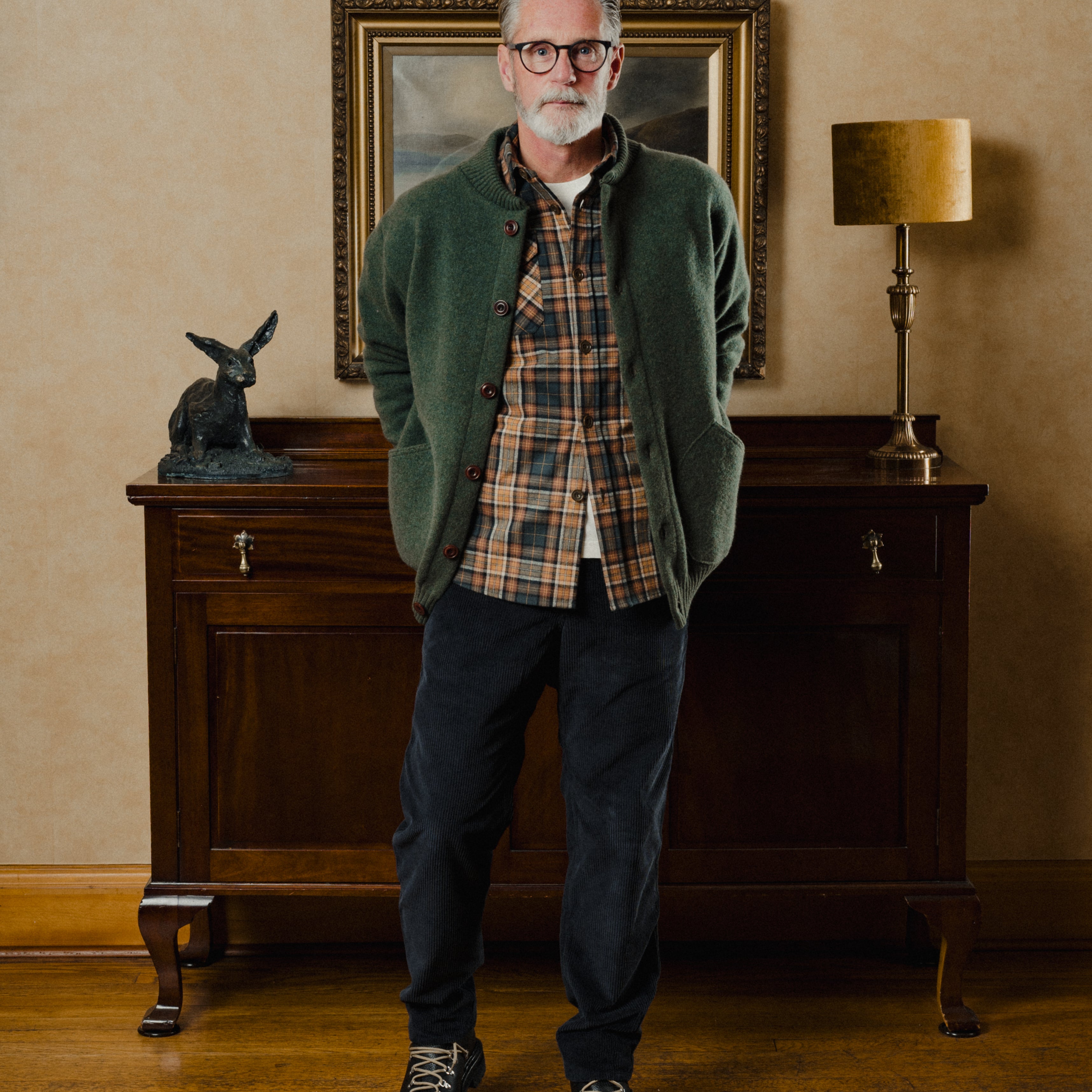 Man wearing a green cardigan over a plaid shirt in a room with a wooden cabinet and lamp.