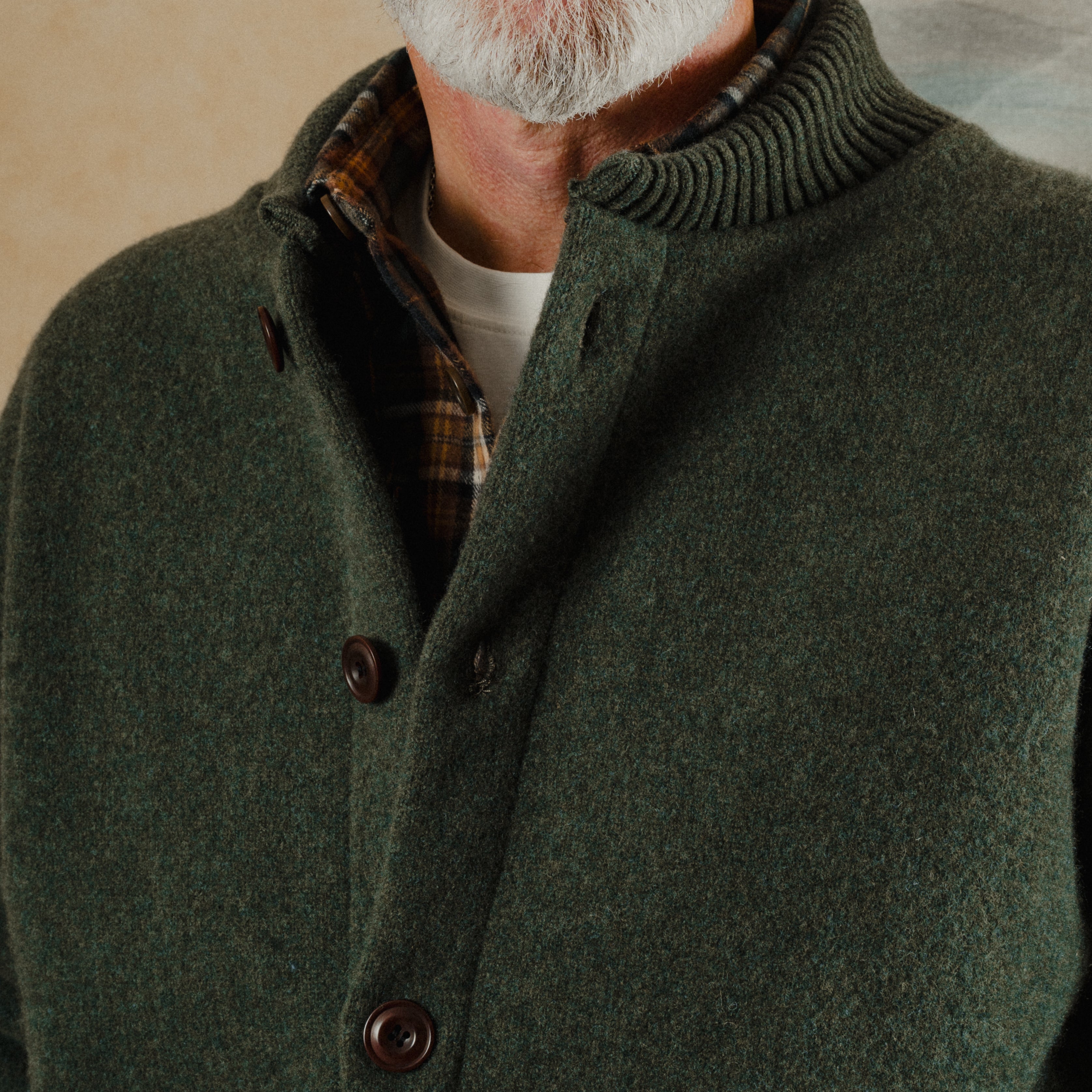 Man wearing a green coat with a plaid shirt underneath, standing against a neutral background.