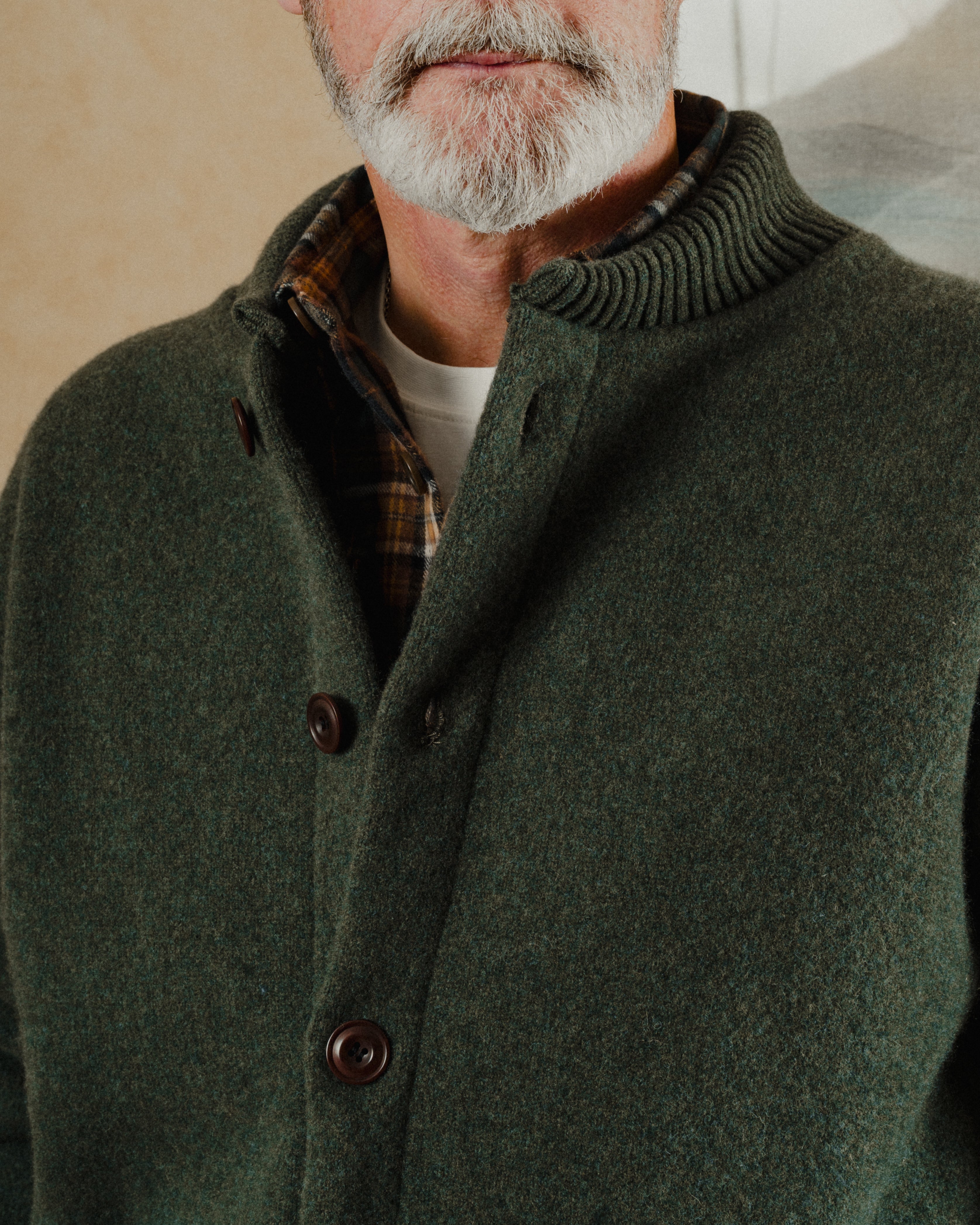 Man wearing a green coat with a plaid shirt underneath, standing against a neutral background.