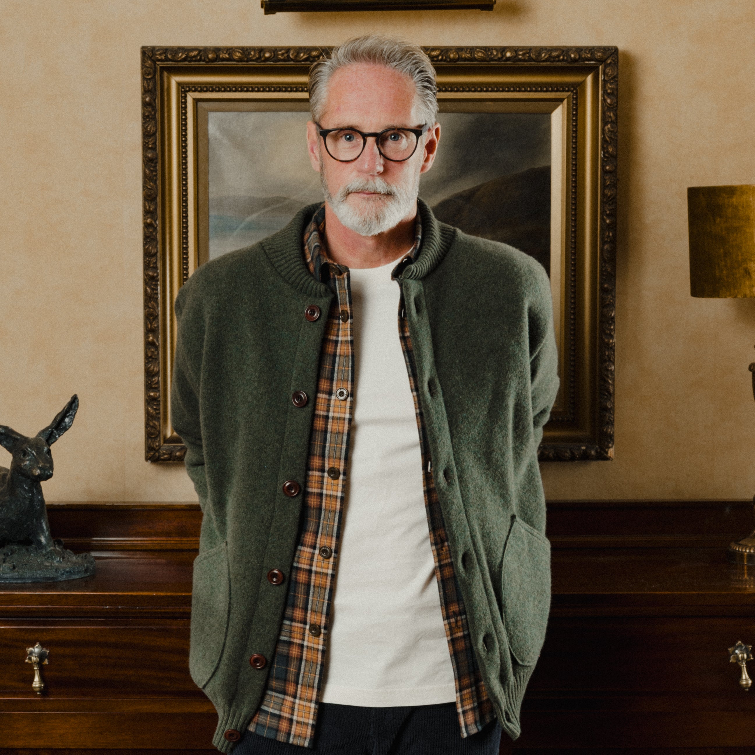 Man wearing glasses and a green jacket standing in a room with a painting on the wall.