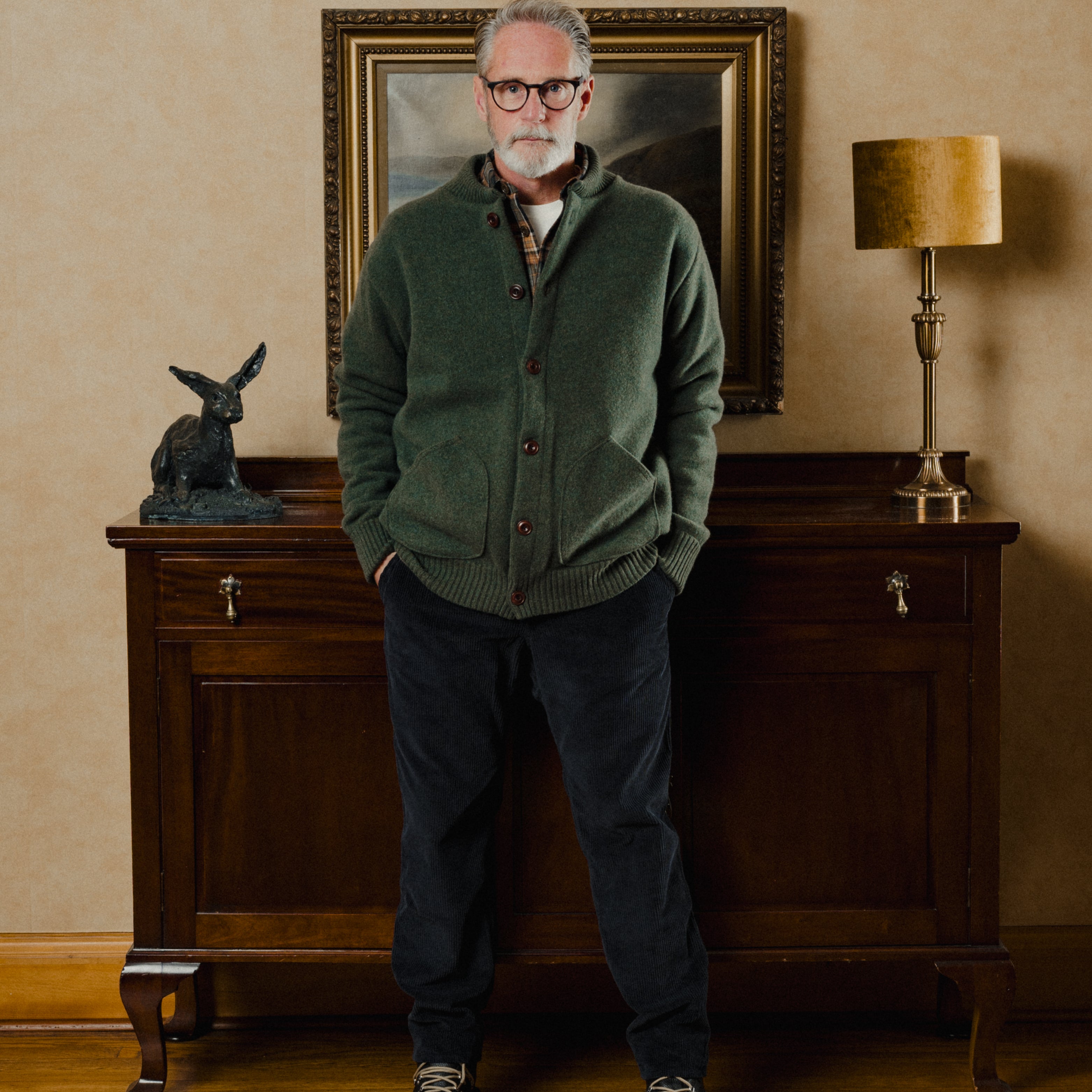 Man wearing a green sweater and dark pants standing in a room with a wooden cabinet and lamp.