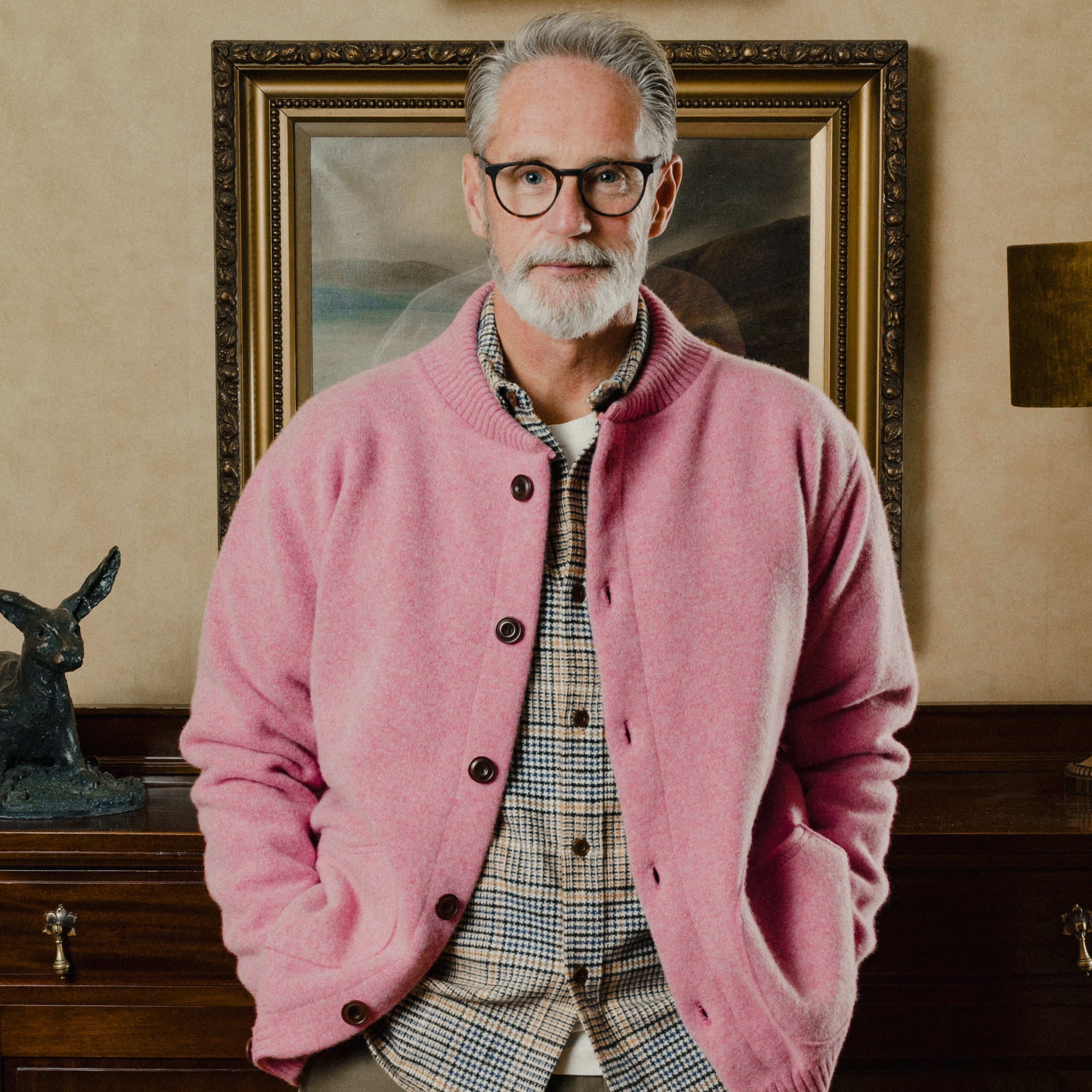 Man wearing a pink cardigan over a plaid shirt in an indoor setting with a painting in the background.