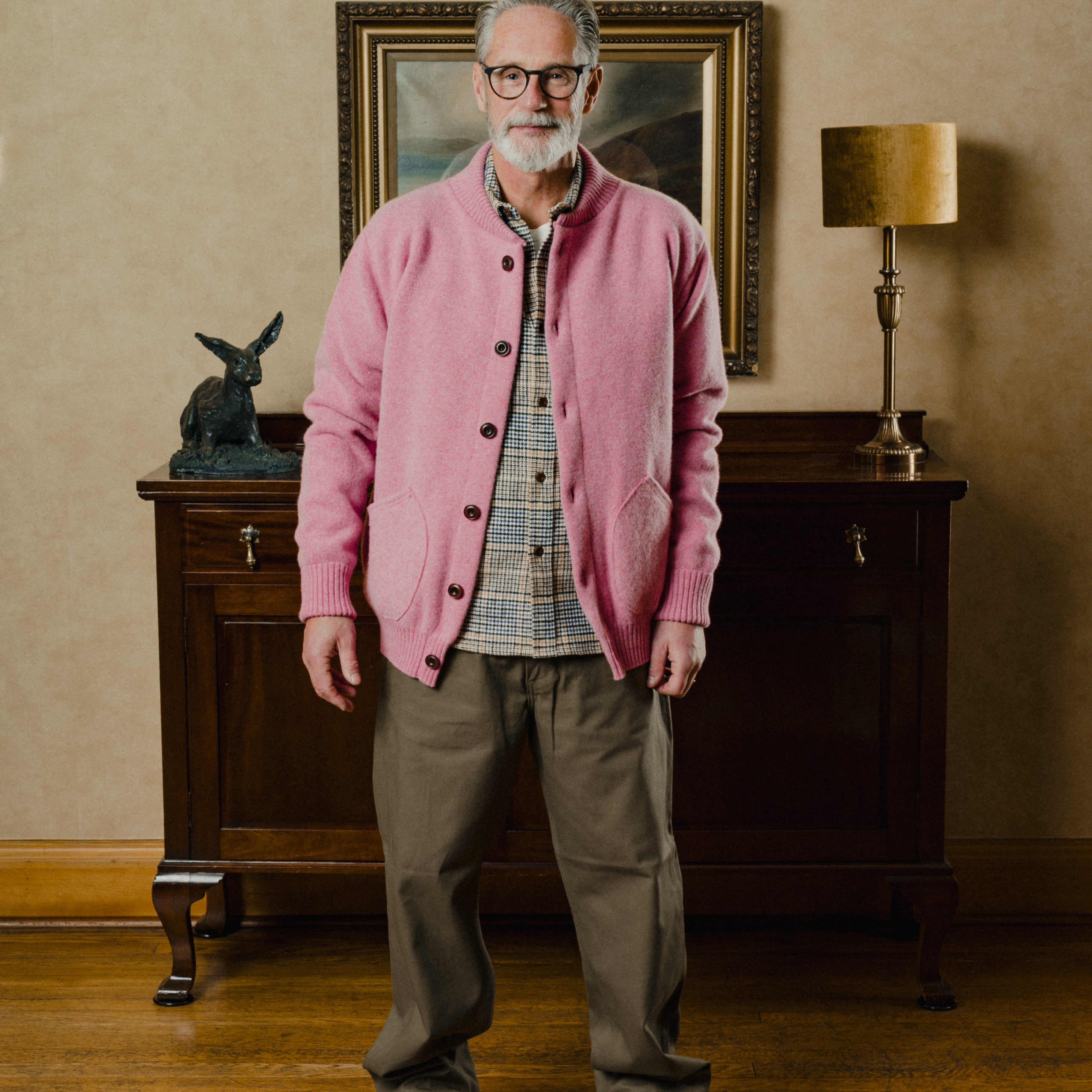 Man wearing a pink cardigan in a room with wooden furniture and a lamp.