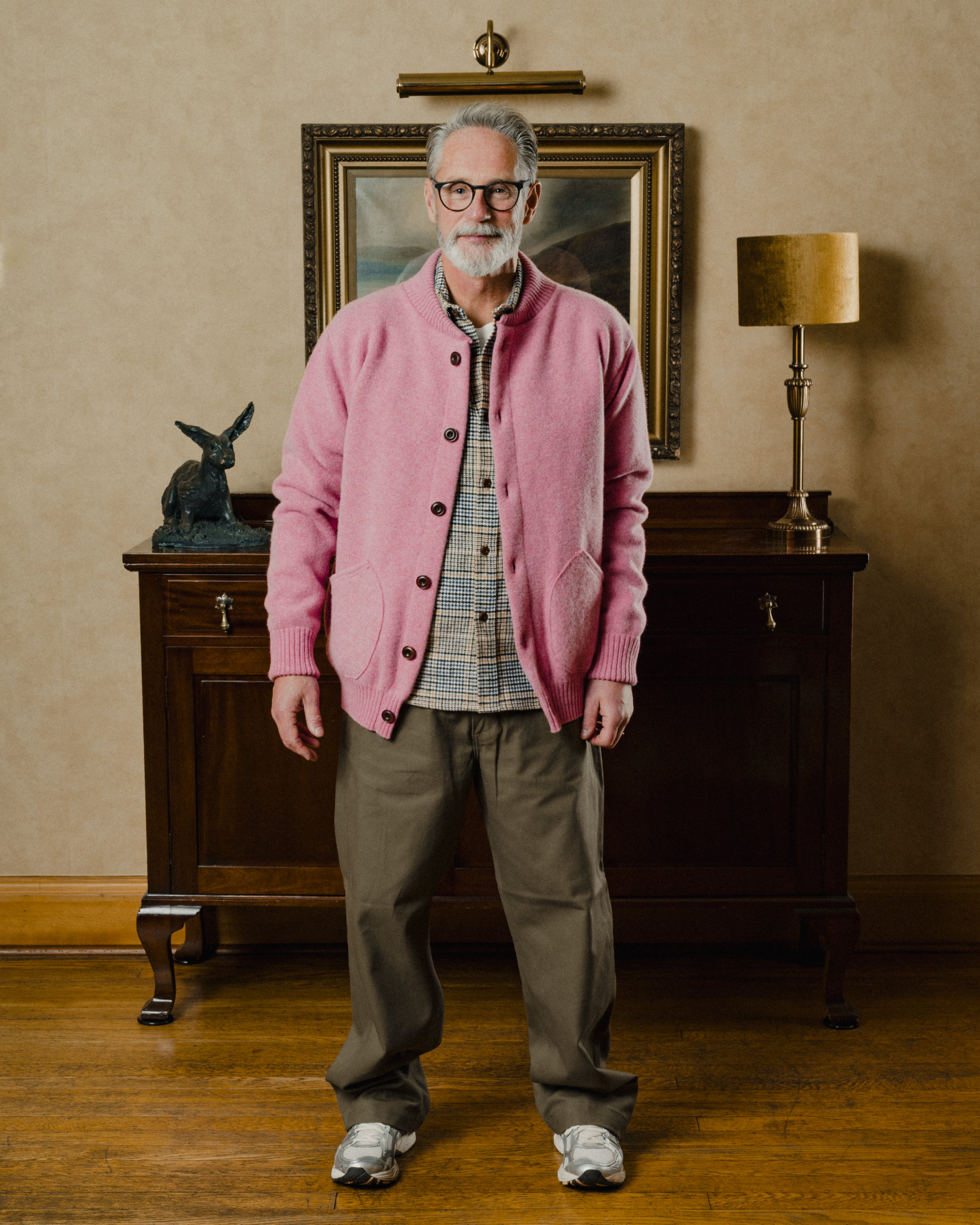 Man wearing a pink cardigan in a room with wooden furniture and a lamp.