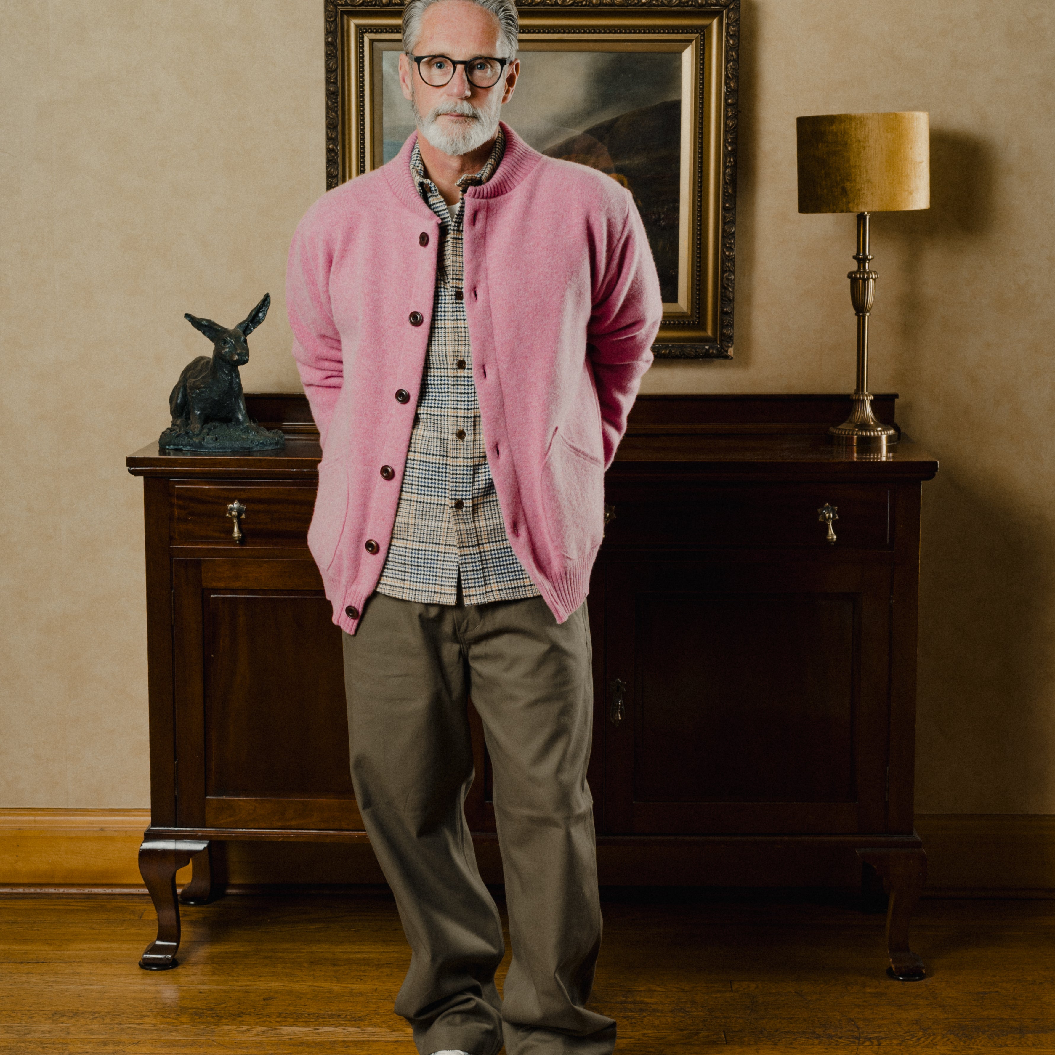 Man wearing a pink cardigan in an elegant room with a painting and lamp.