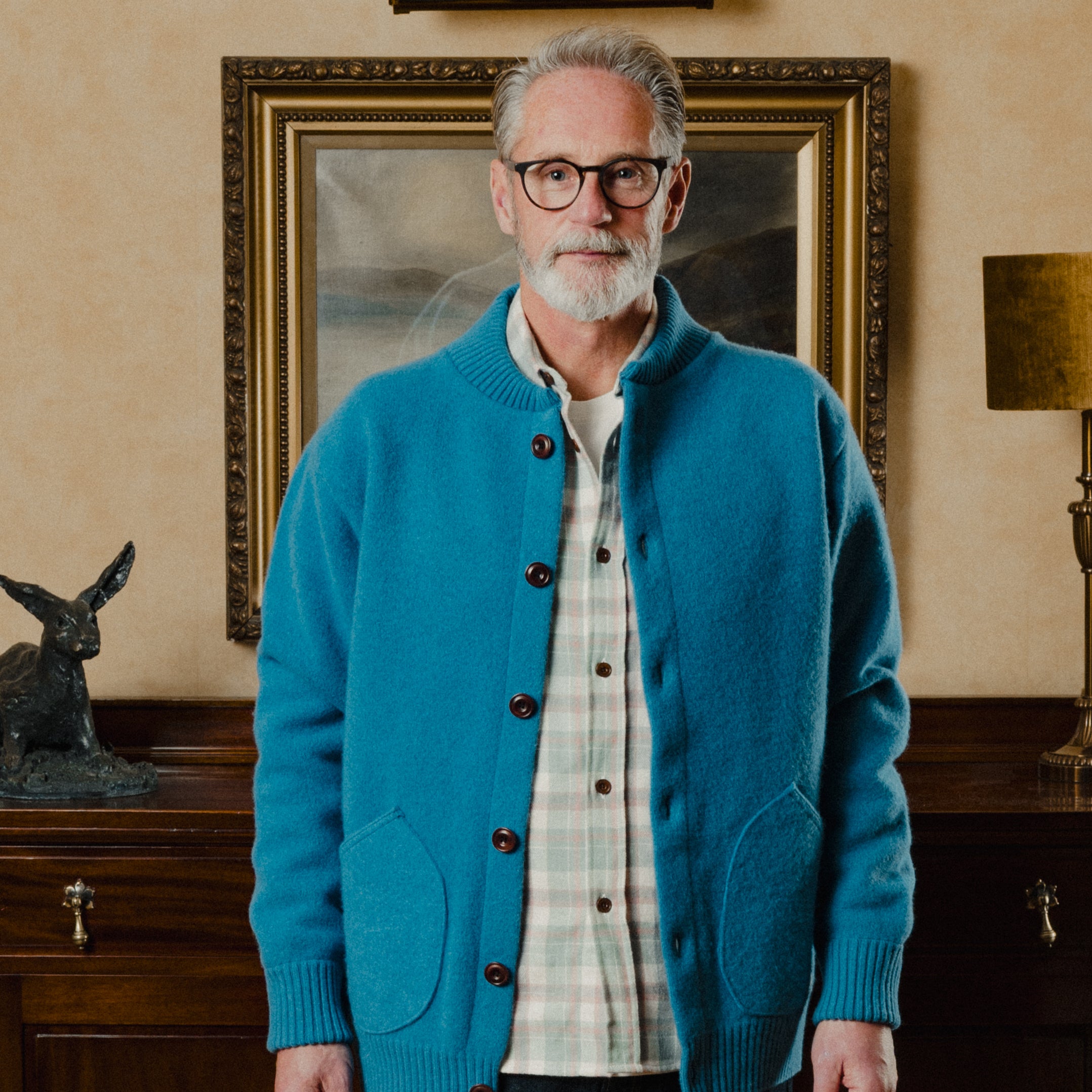 Man wearing a blue cardigan in a room with a painting and lamp in the background