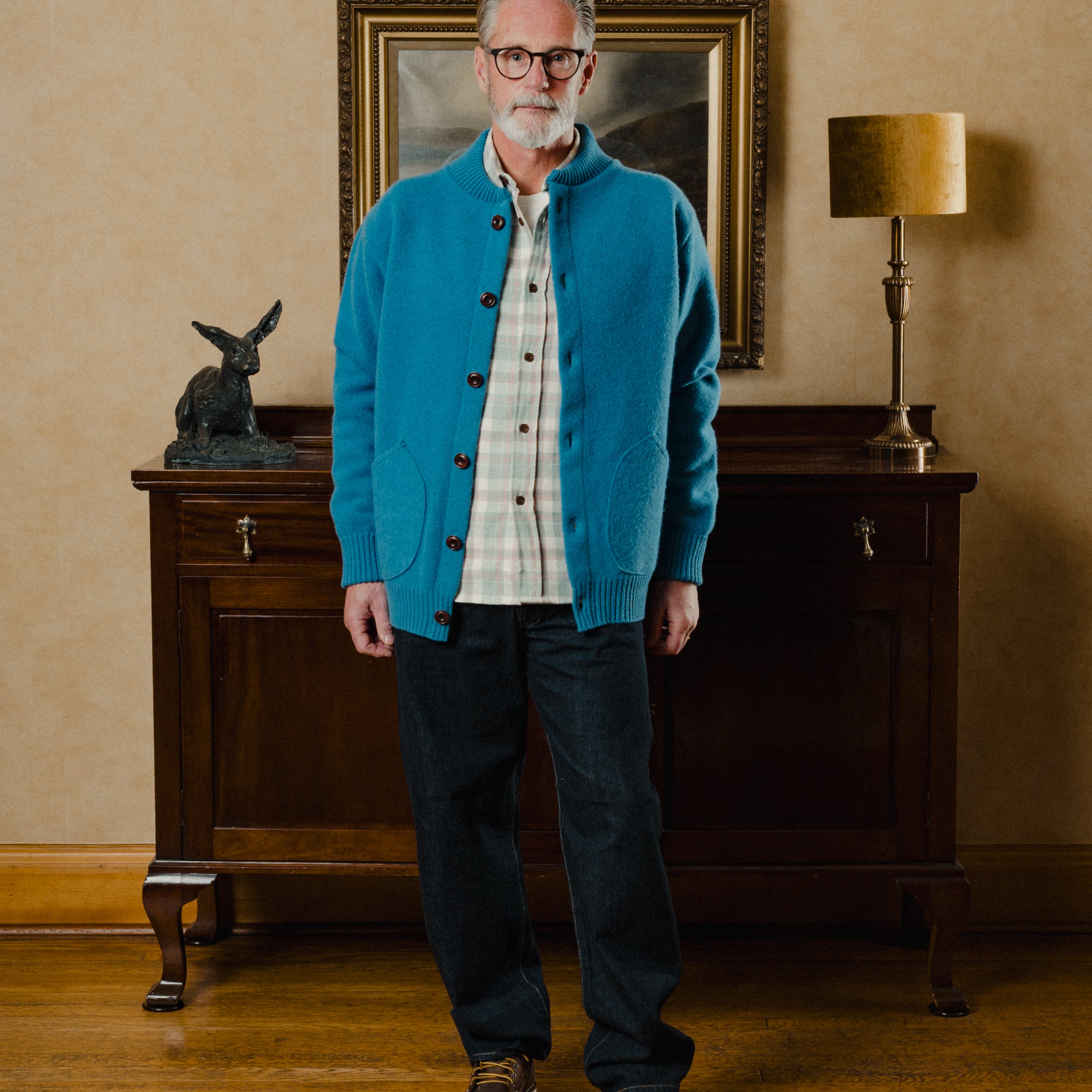 Man wearing a blue cardigan in an indoor setting with a lamp and painting in the background.