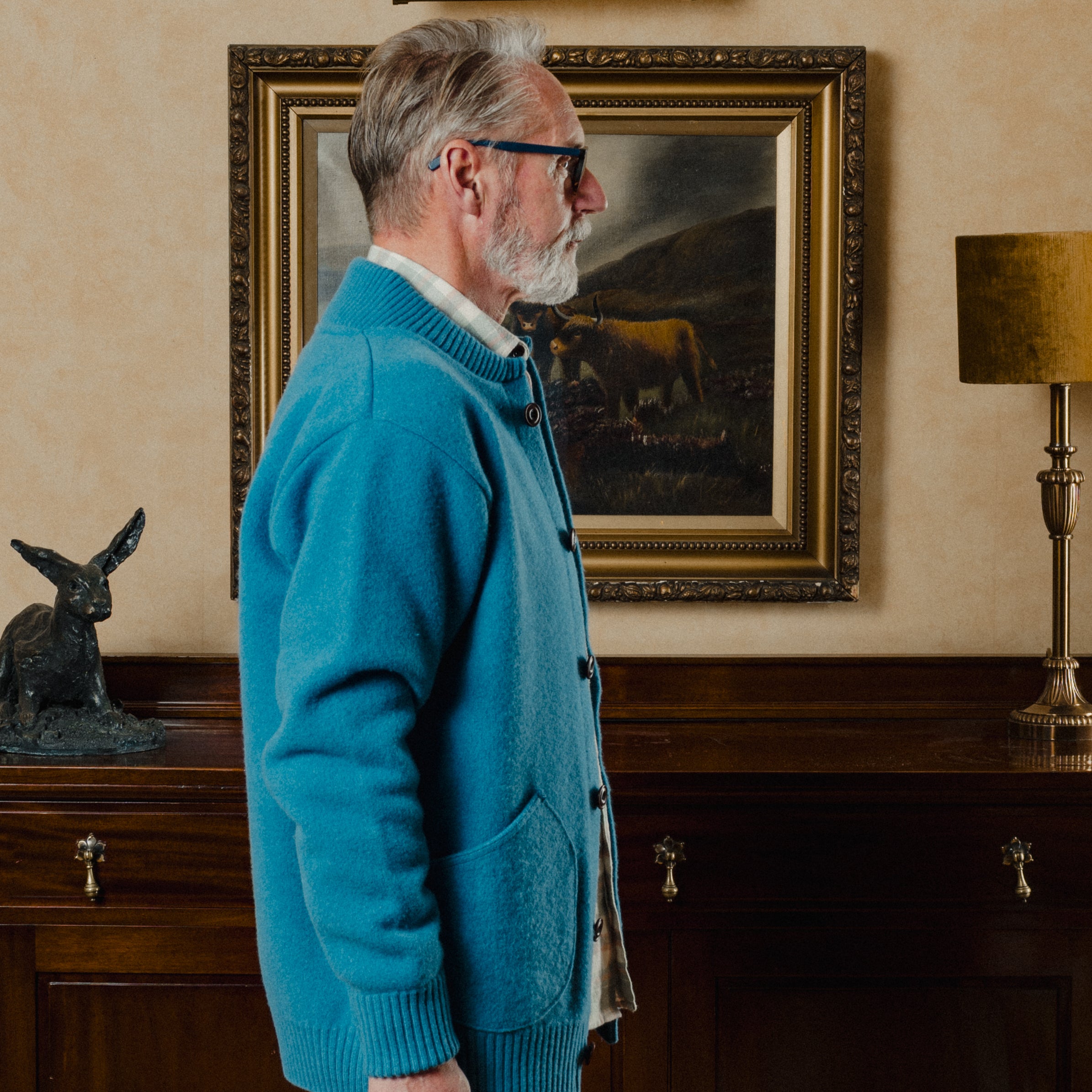 Man in a blue cardigan standing in a room with a painting of a cow on the wall.
