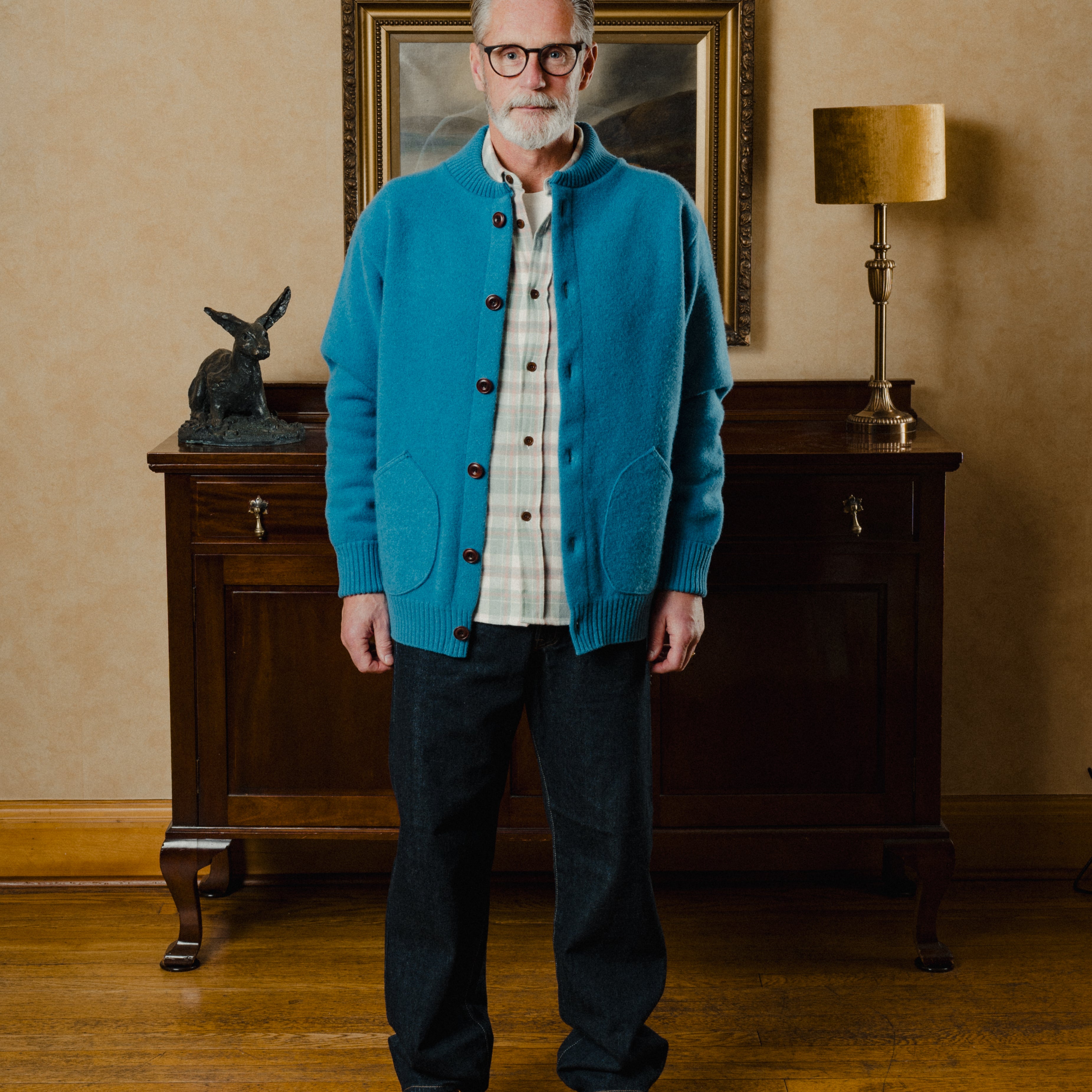 Man wearing a blue cardigan in a room with wooden furniture and a lamp.