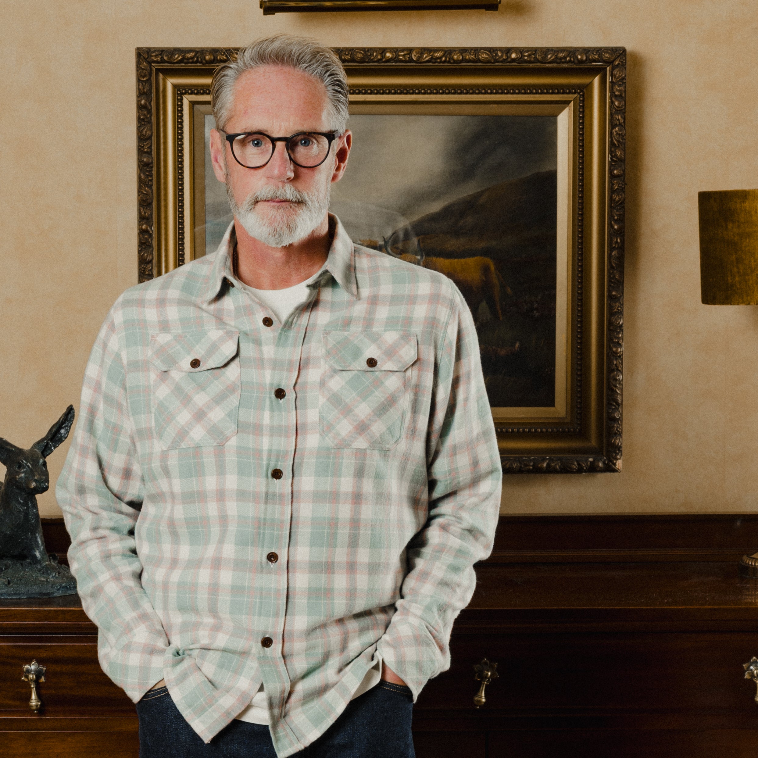 Man wearing a plaid shirt standing in front of a framed painting on a wall.