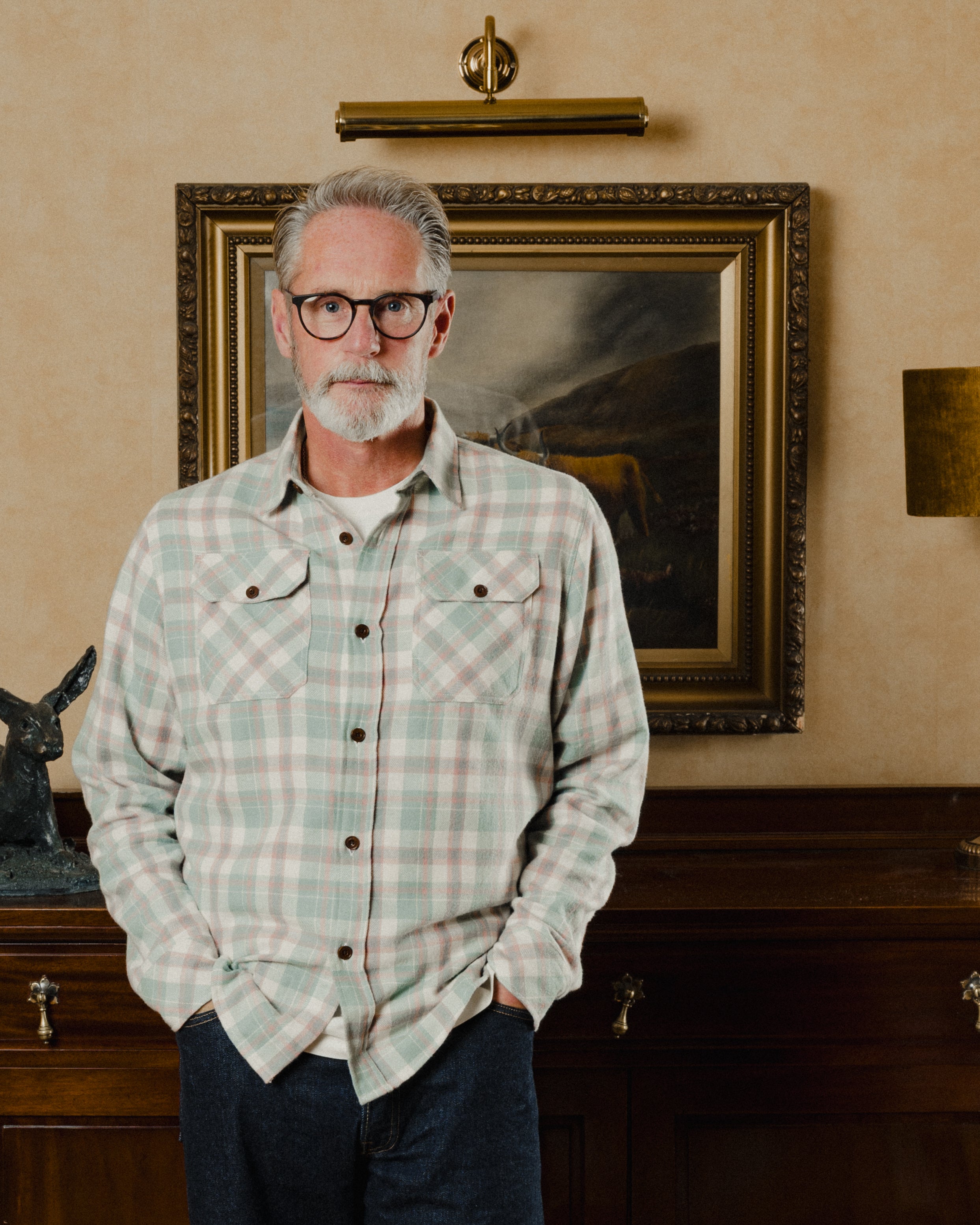 Man wearing a plaid shirt standing in front of a framed painting on a wall.