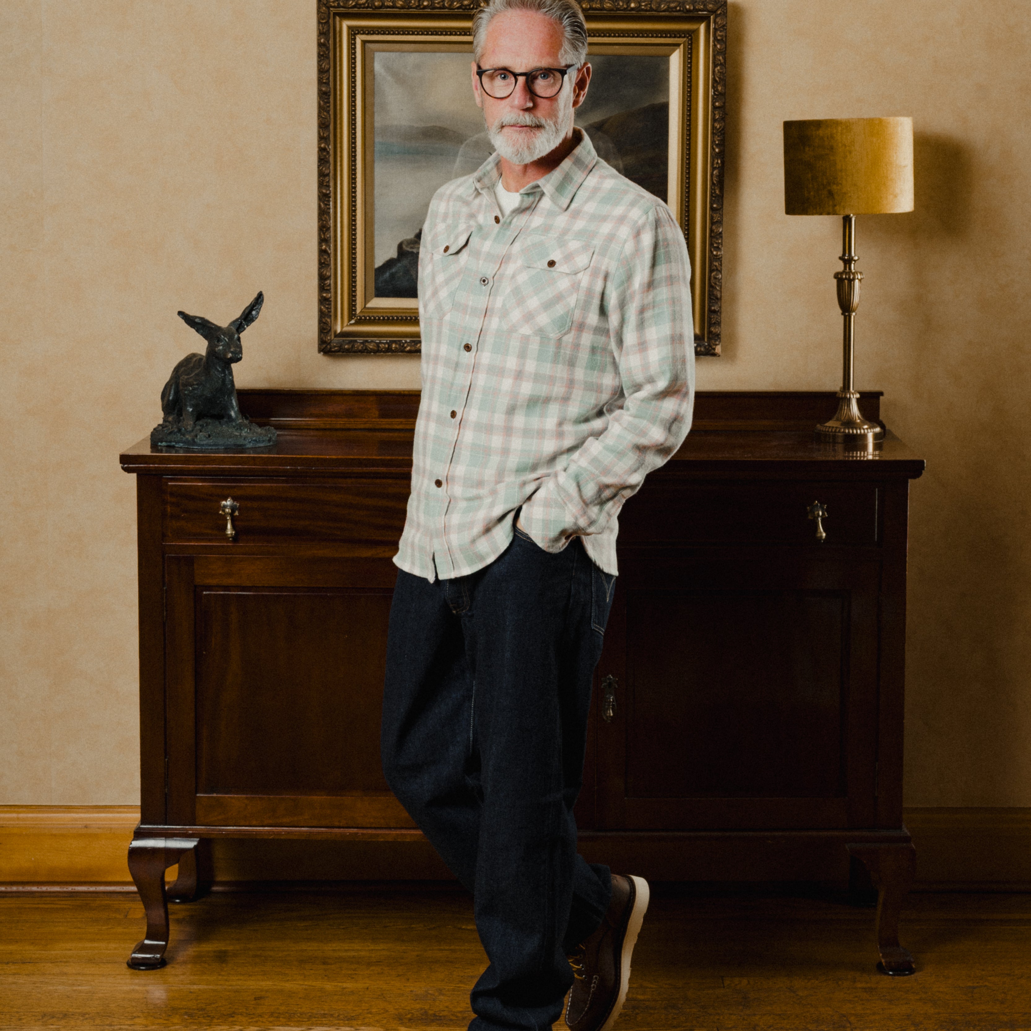 Man standing in a room with a painting on the wall and a lamp on a cabinet.