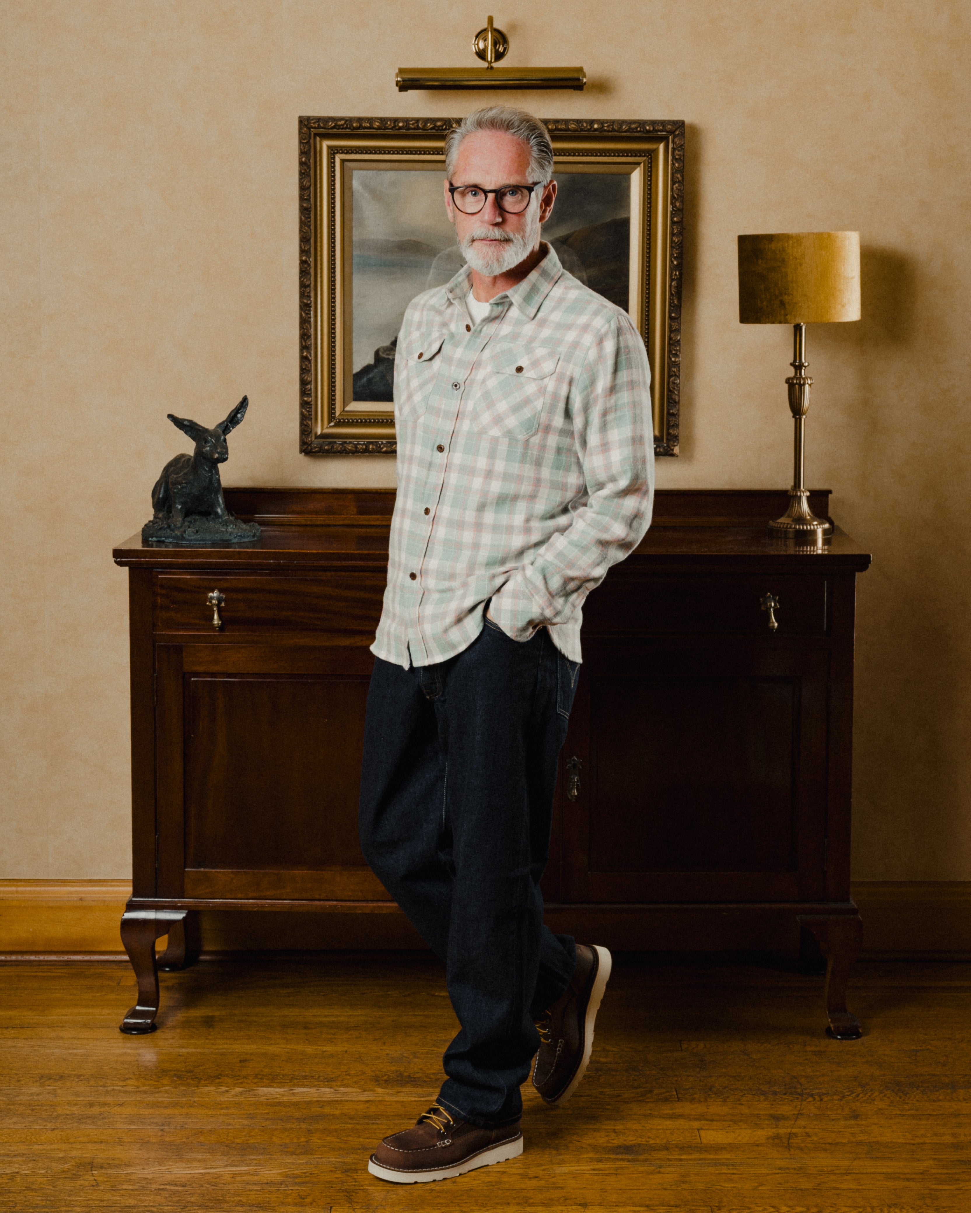 Man standing in a room with a painting on the wall and a lamp on a cabinet.