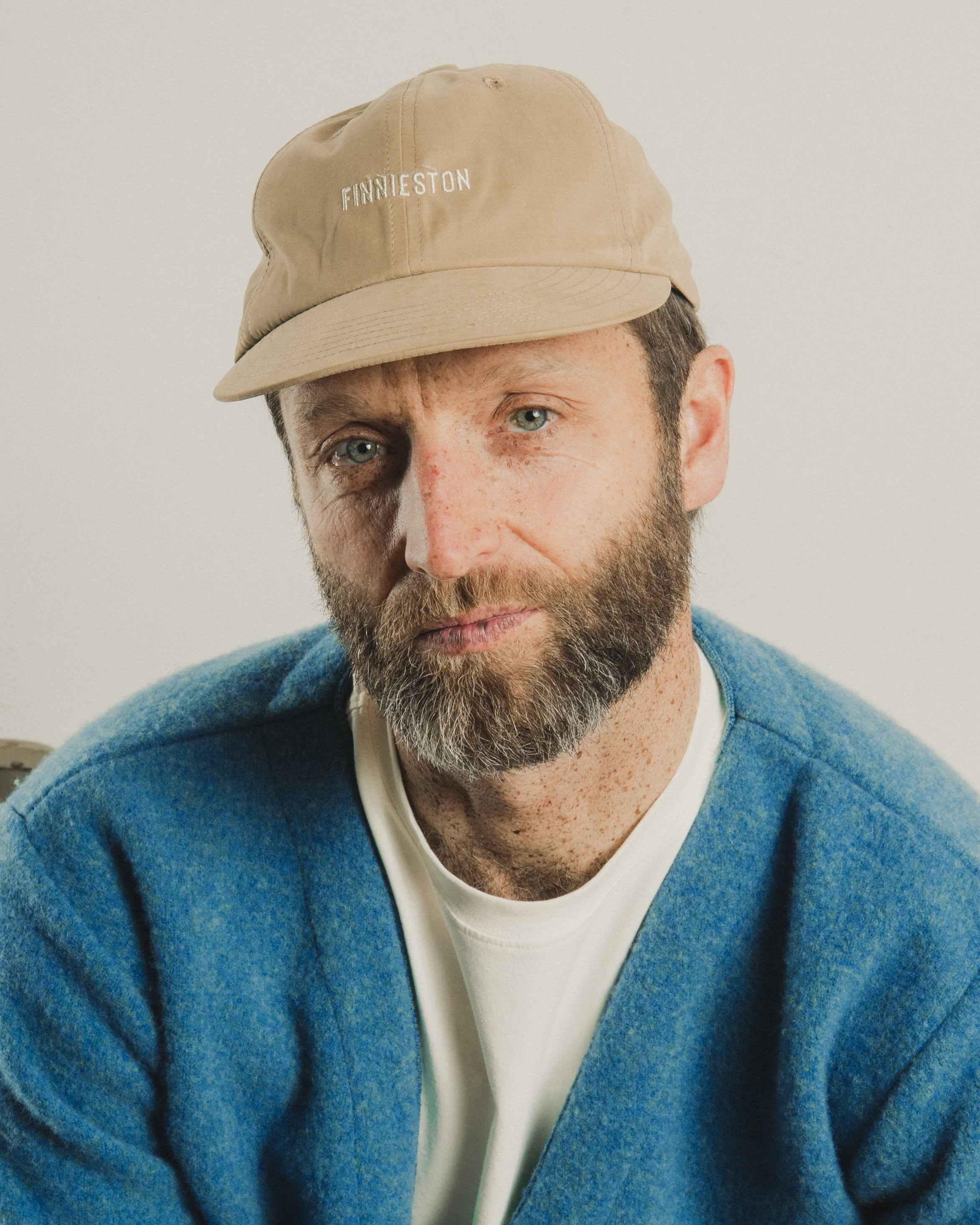 Man wearing a beige cap with 'FINNION' branding, blue cardigan, and white shirt against a plain background
