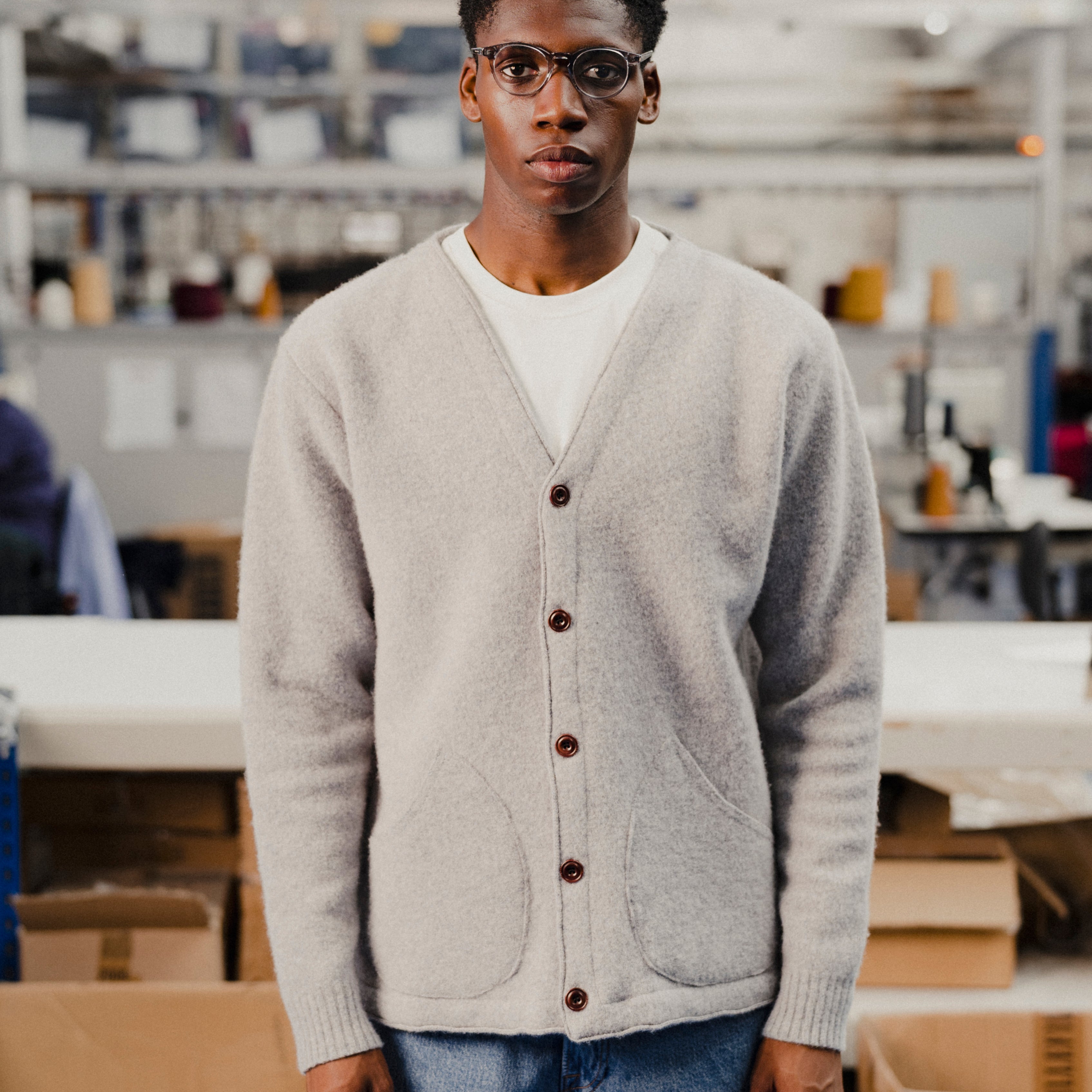 Man wearing a grey cardigan in an indoor setting with shelves and boxes in the background