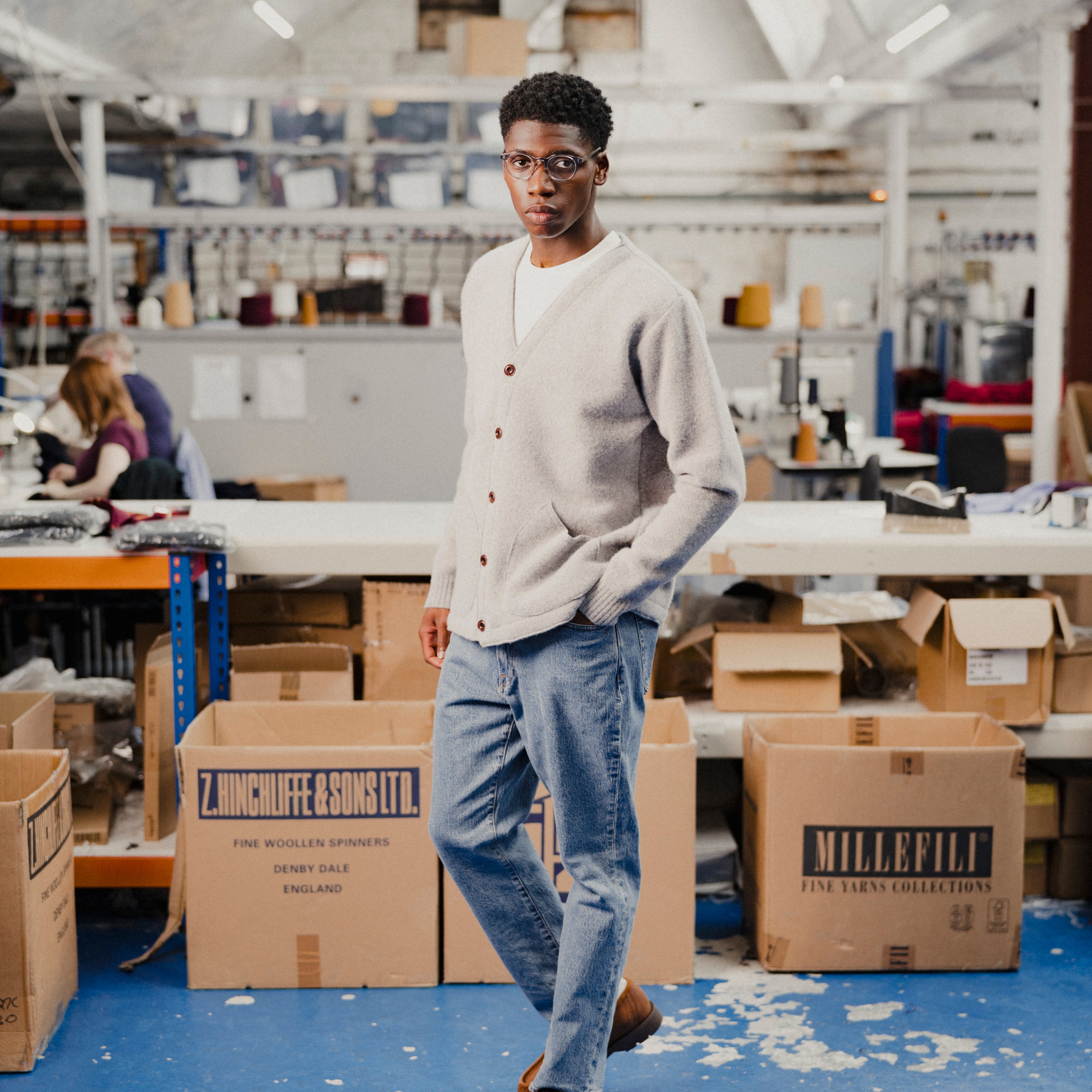 Man wearing a light grey cardigan in a fashion warehouse, stood around boxes