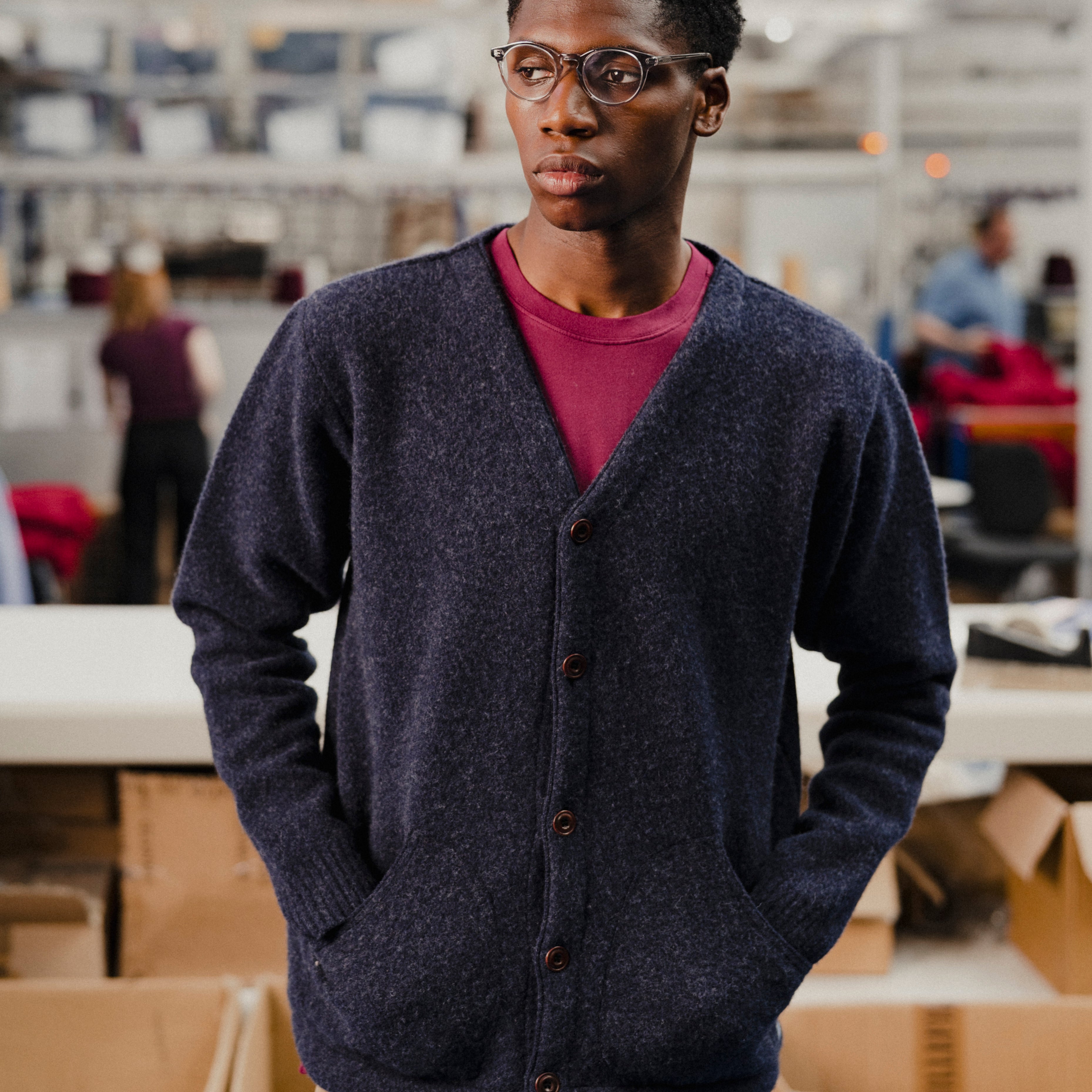 Person wearing a dark blue cardigan over a red shirt in an indoor setting with boxes and people in the background.