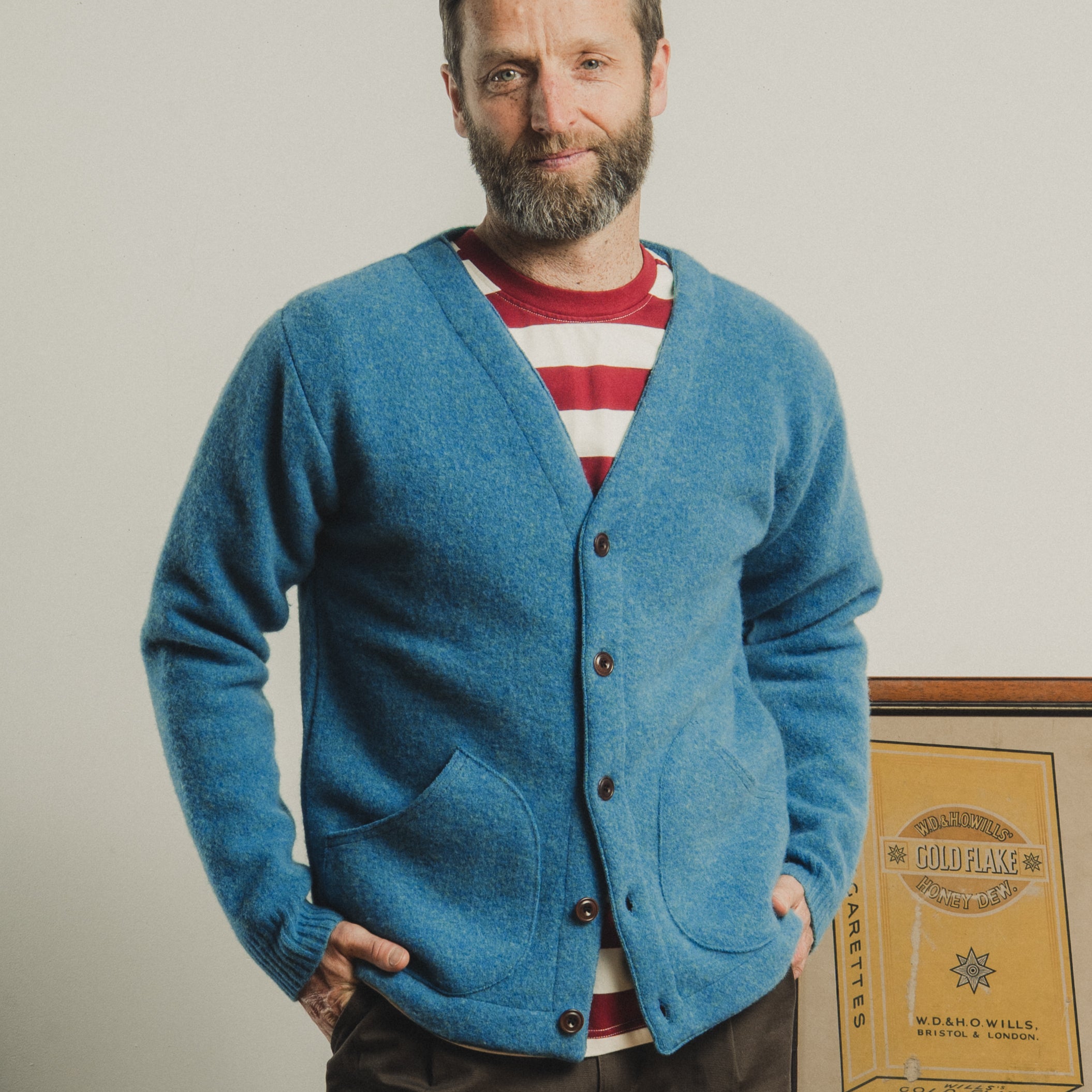 Man wearing a blue cardigan over a red and white striped shirt, standing against a plain background.
