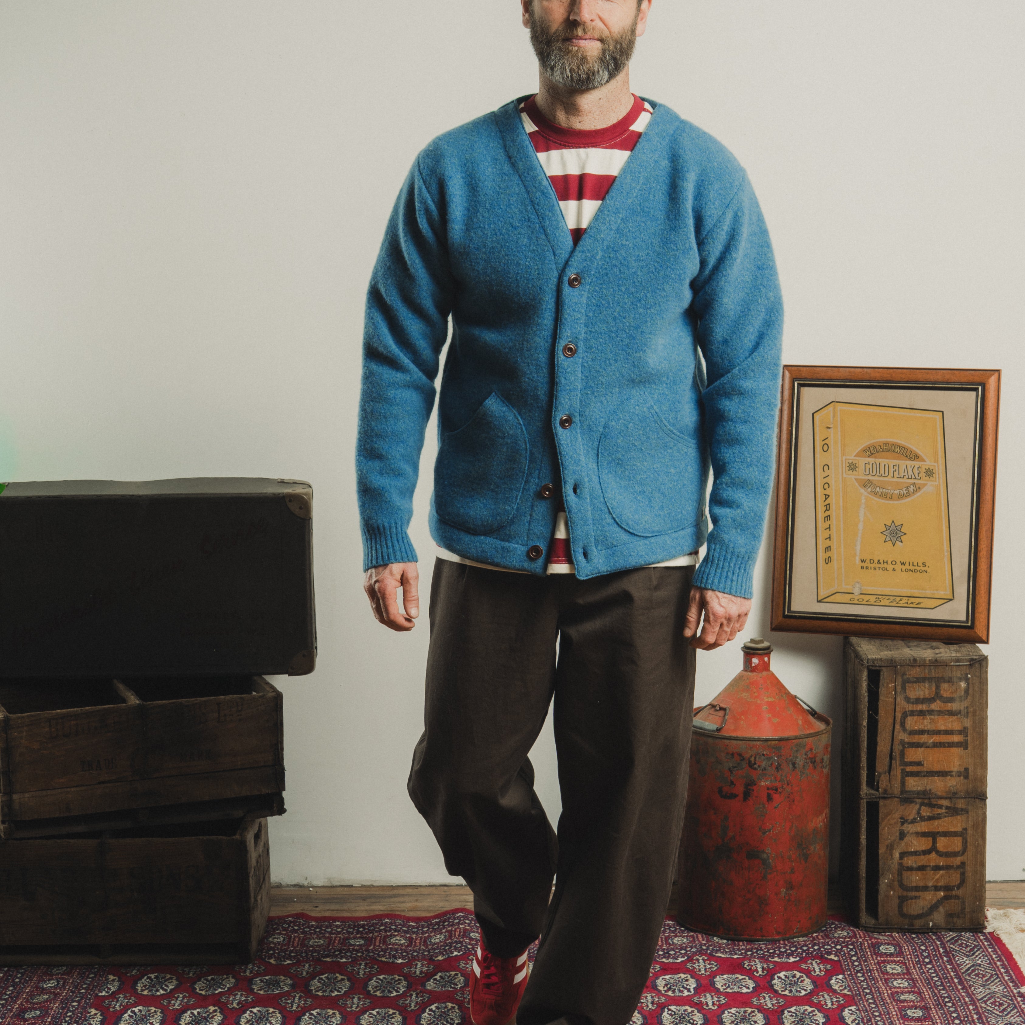 Man wearing a blue cardigan over a striped shirt, standing in a room with wooden crates and a red fire extinguisher.