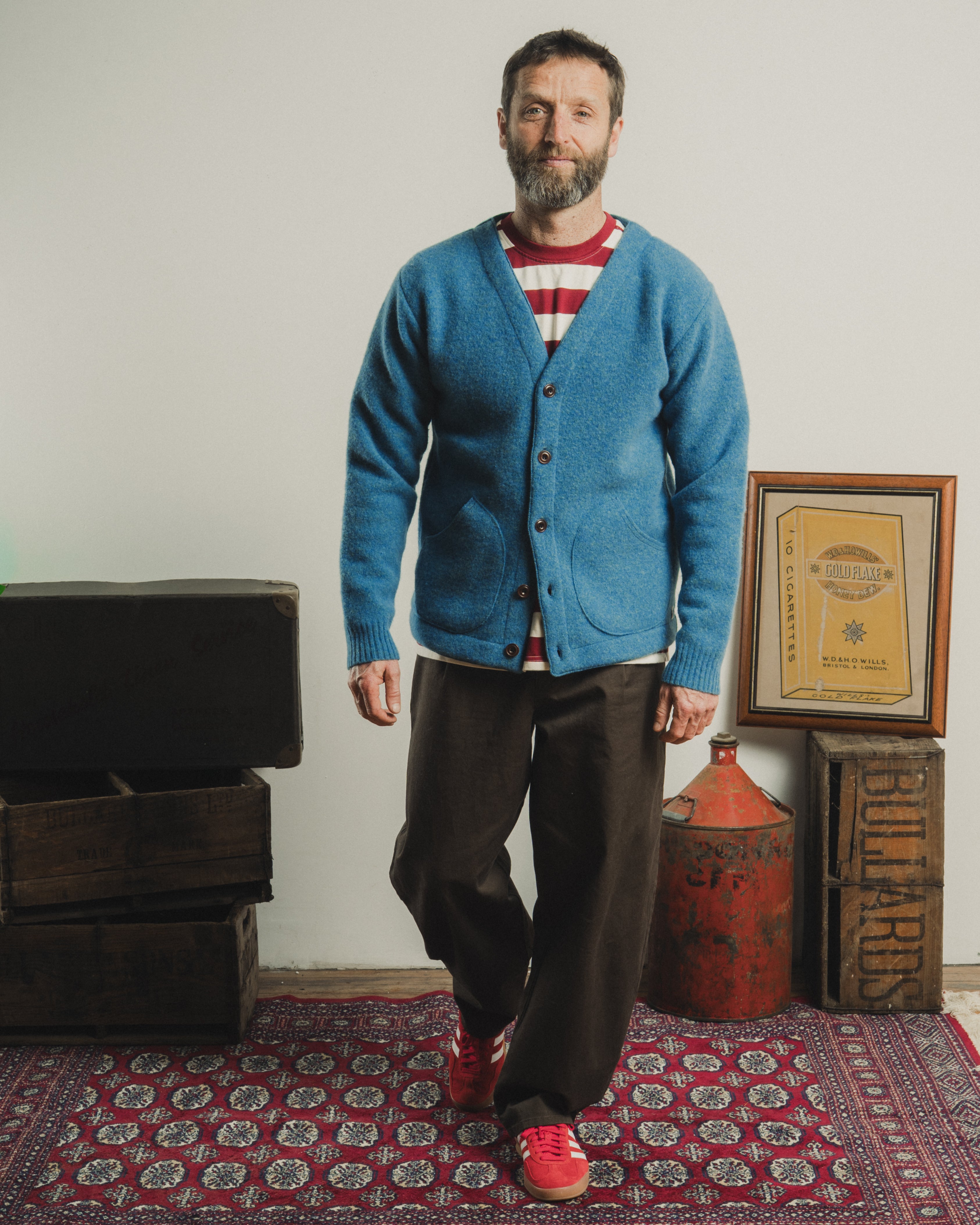 Man wearing a blue cardigan over a striped shirt, standing in a room with wooden crates and a red fire extinguisher.