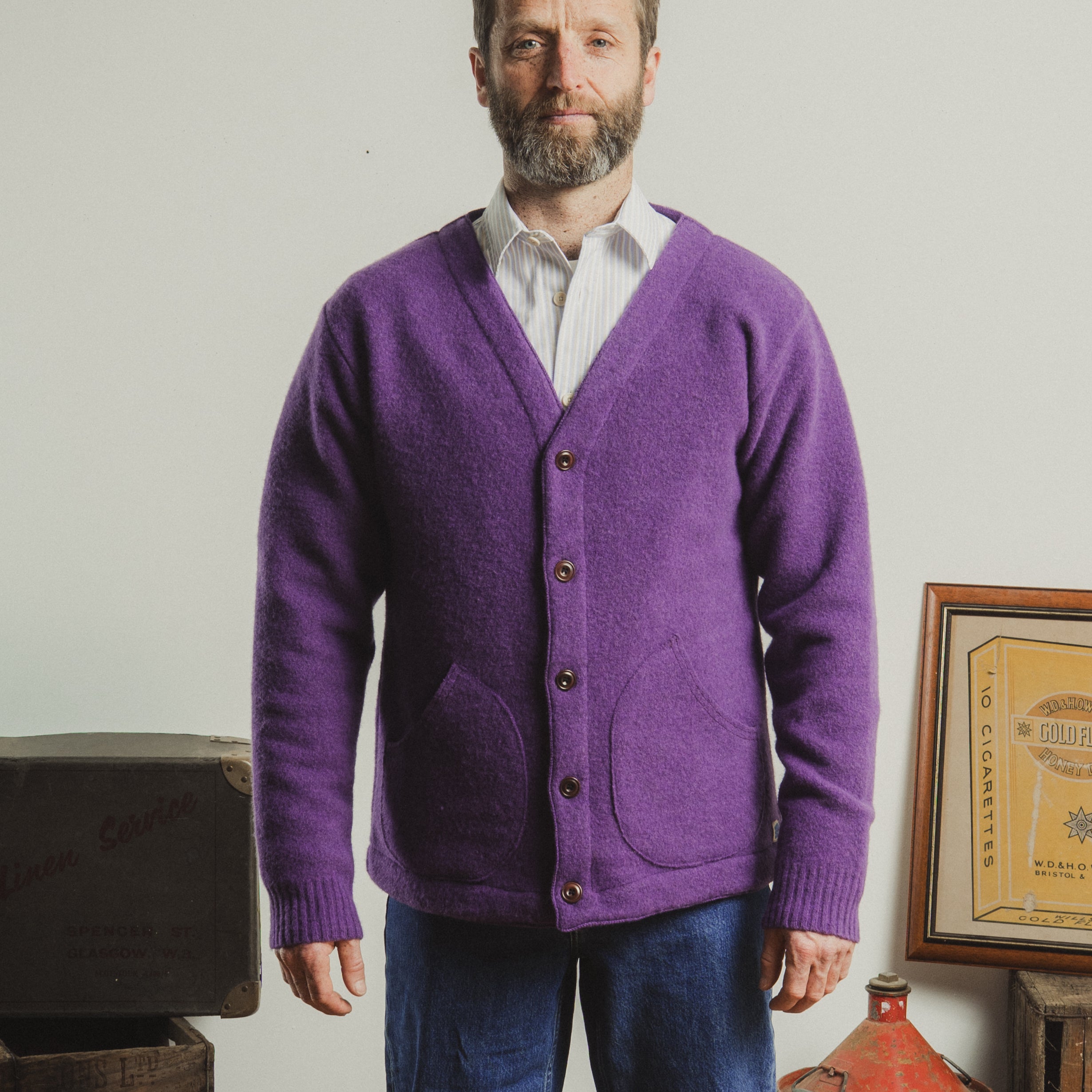 Man wearing a purple cardigan over a white shirt, standing in a room with a plain background.