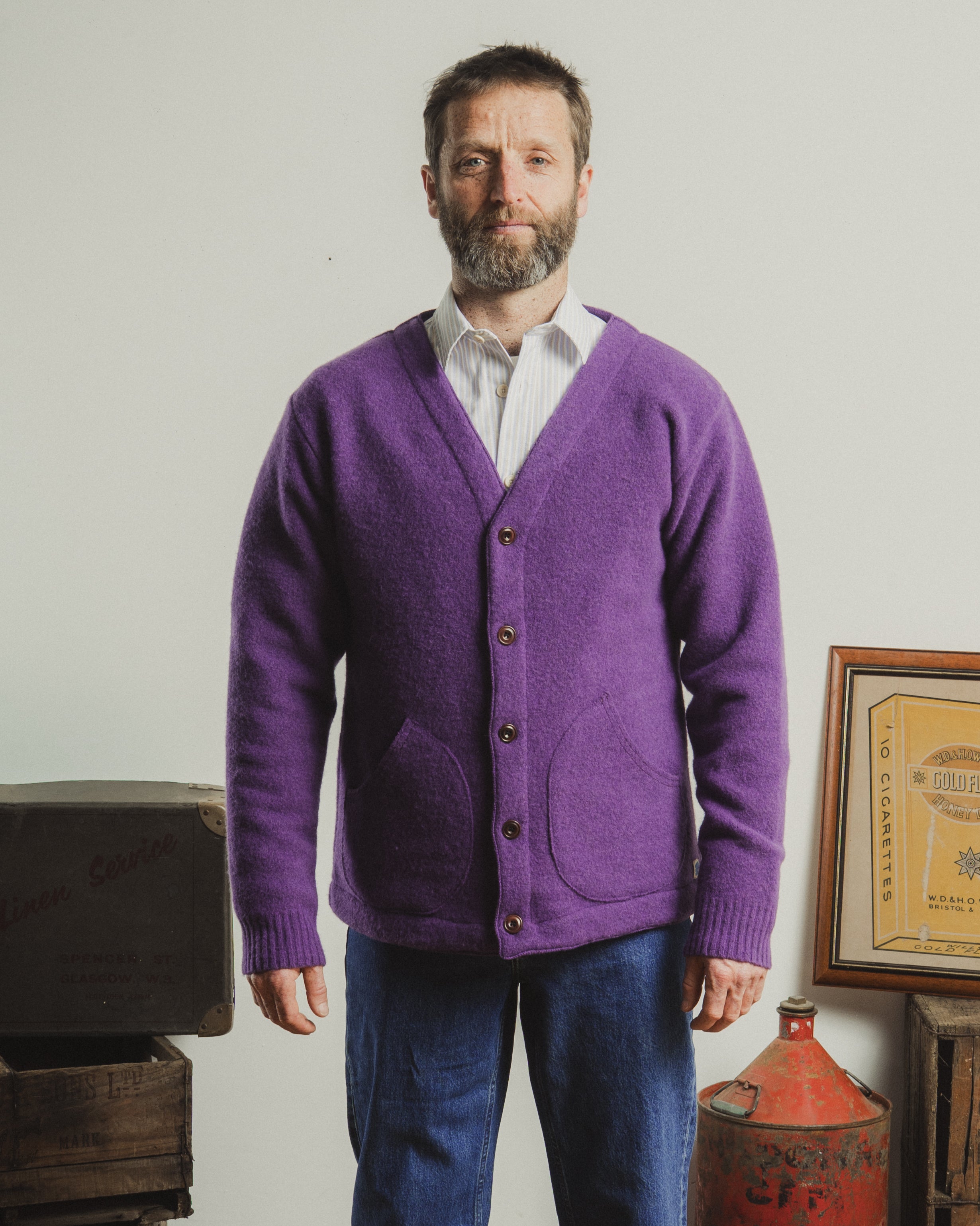 Man wearing a purple cardigan over a white shirt, standing in a room with a plain background.