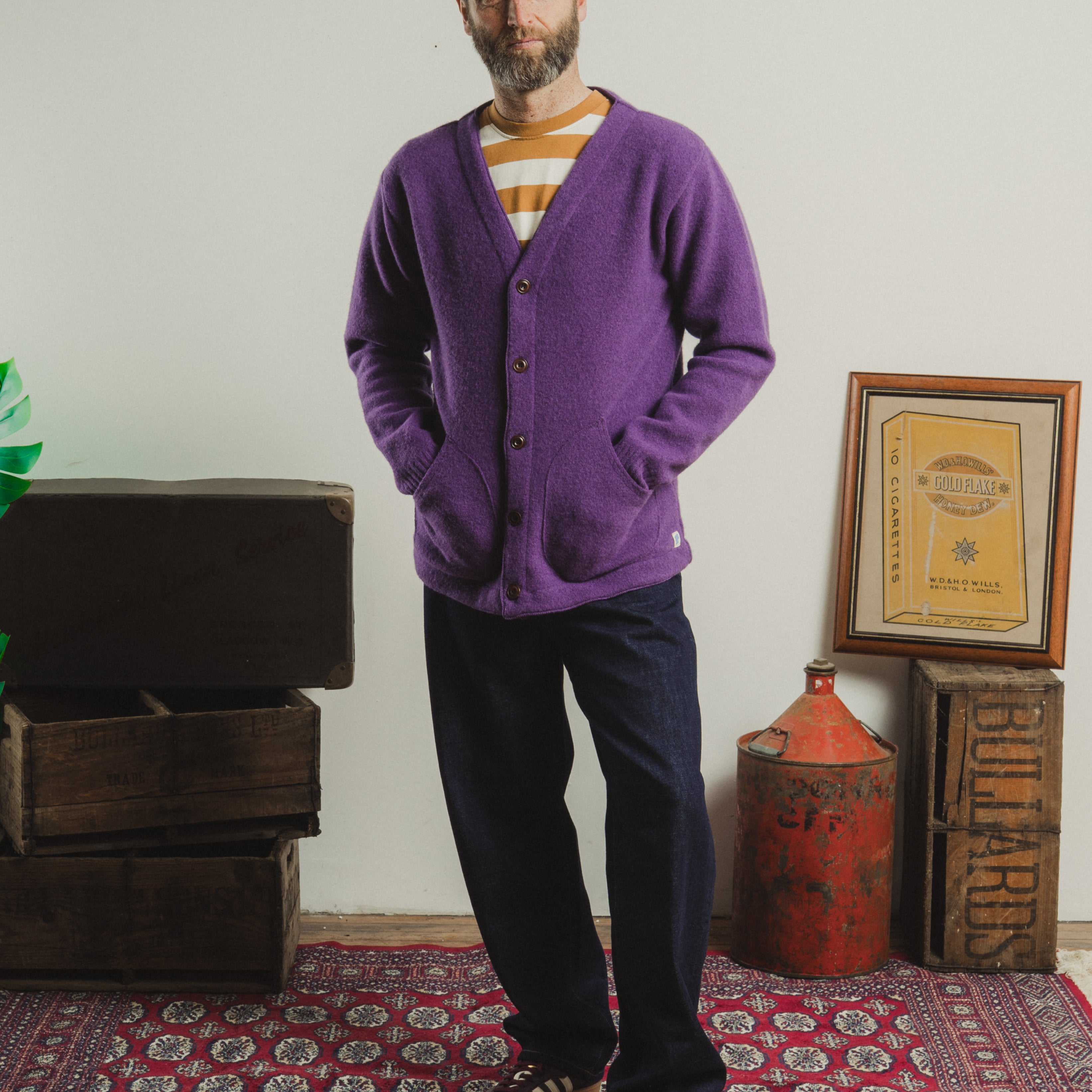 Man wearing a purple cardigan standing in a room with a patterned rug and rustic decor.