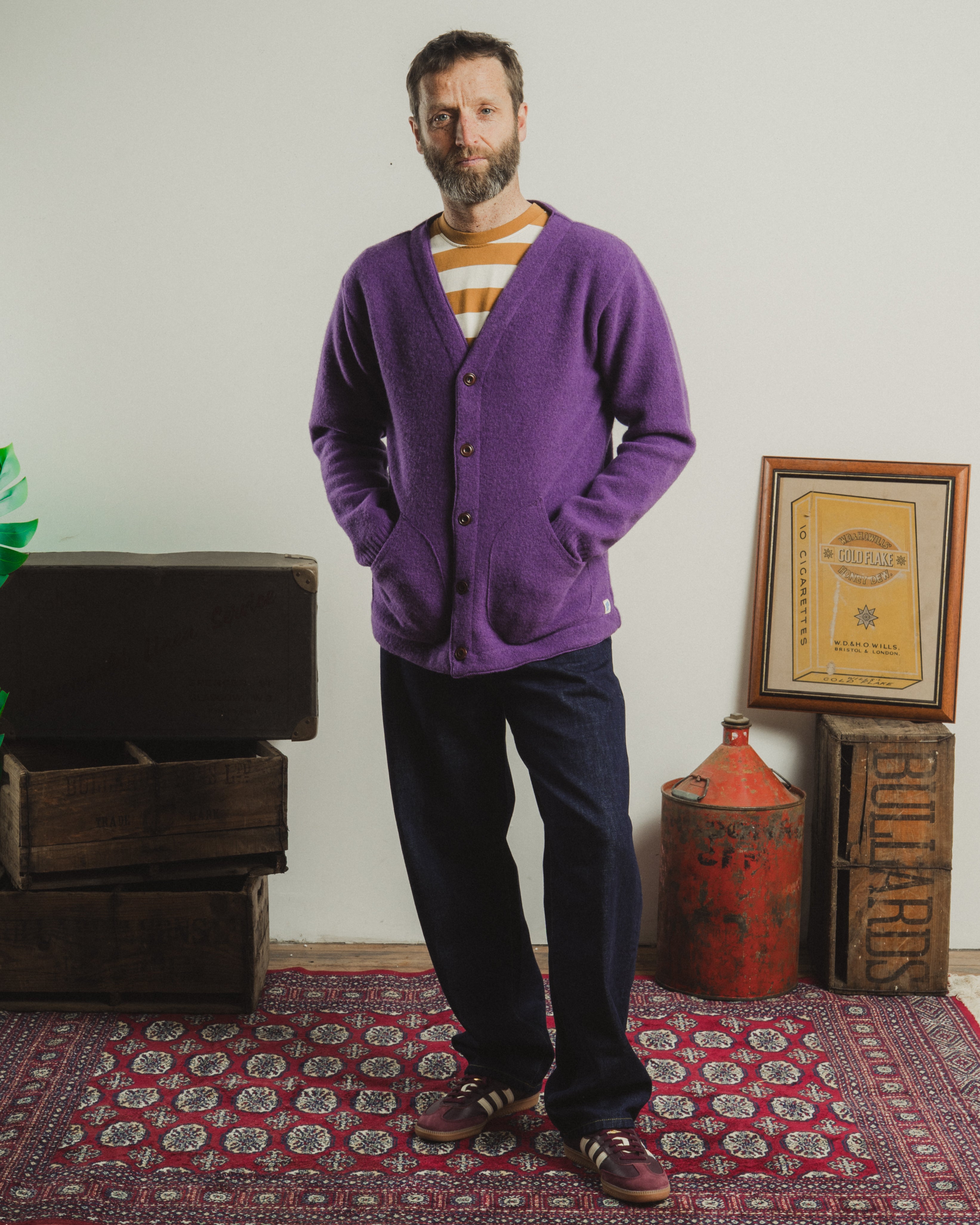 Man wearing a purple cardigan standing in a room with a patterned rug and rustic decor.