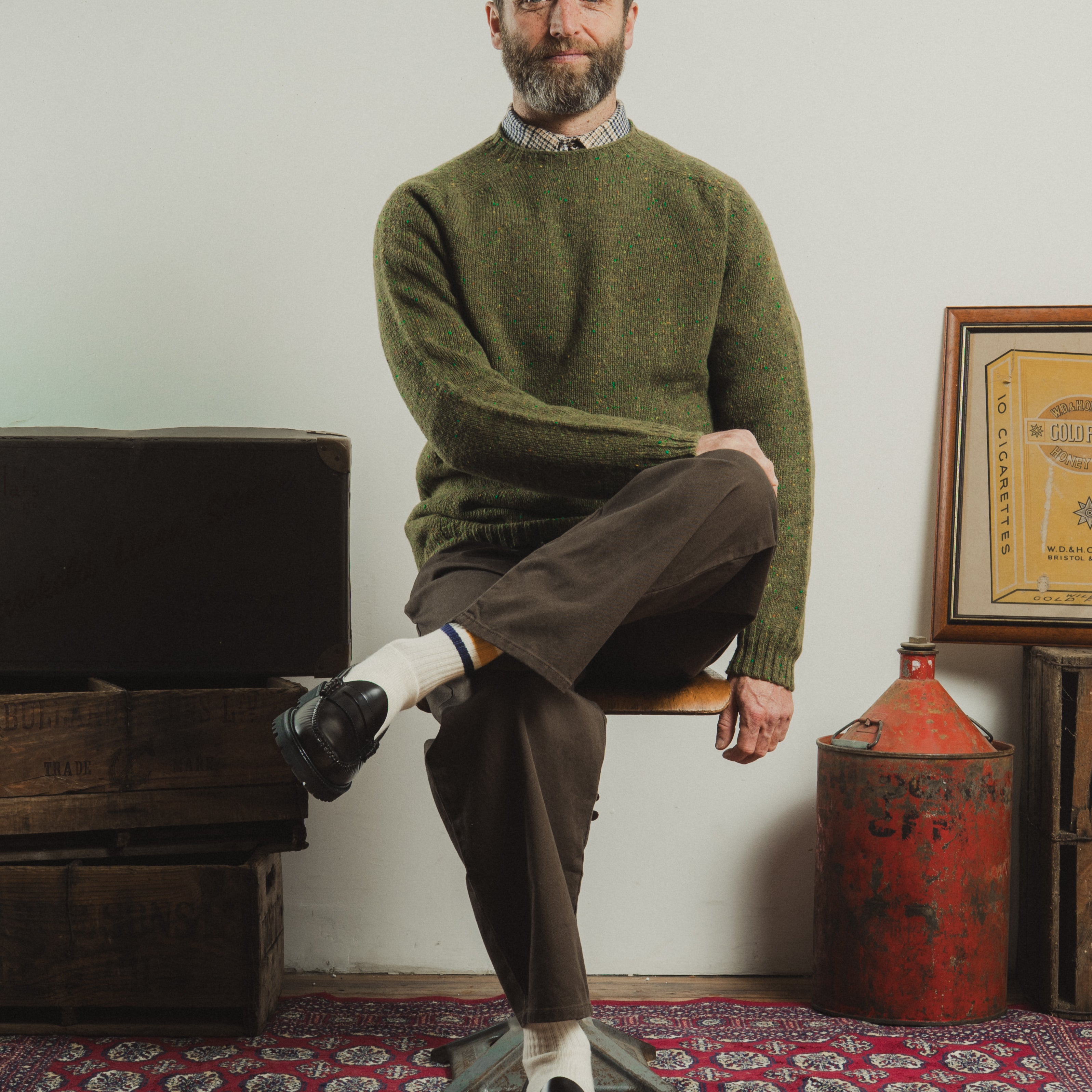 Man sitting on a chair wearing a green sweater and brown pants in a room with rustic decor.