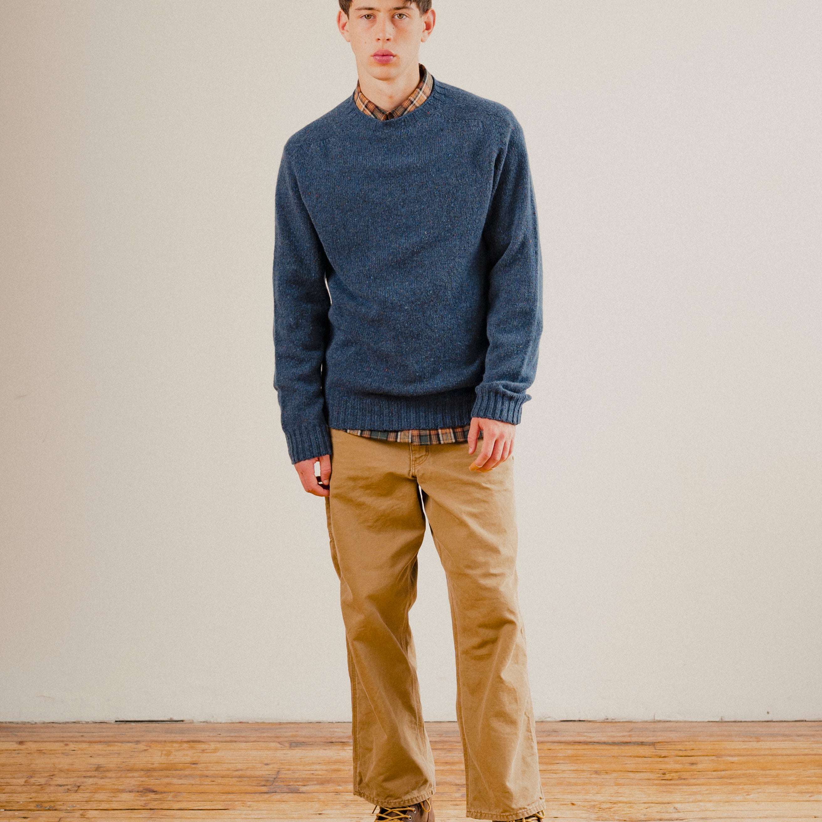 Man wearing a blue wool sweater knit crewneck with a check shirt underneath on a plain background with a wooden floor