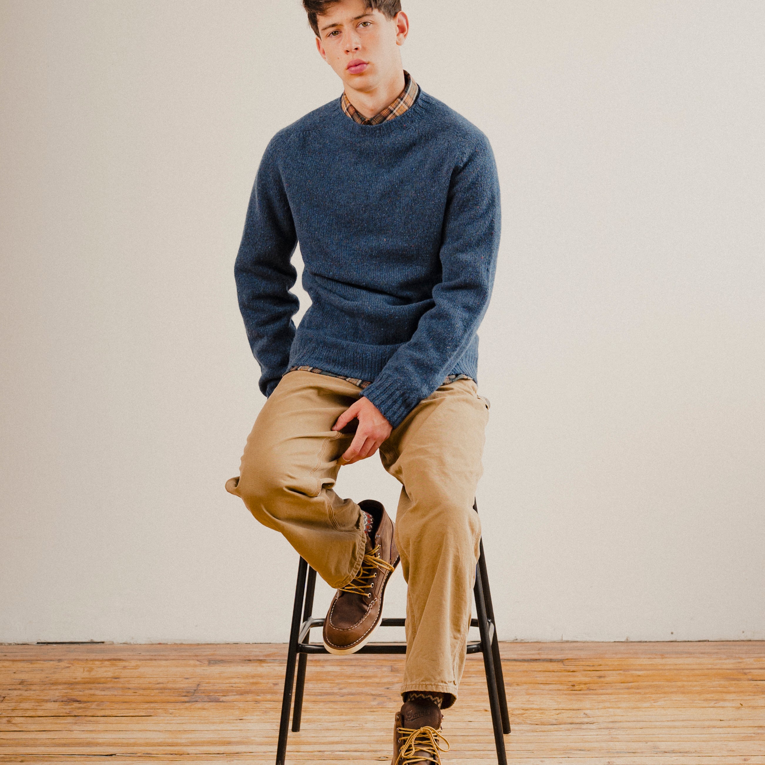 Man sat on a stool wearing a navy blue wool sweater knit crewneck with a check shirt underneath