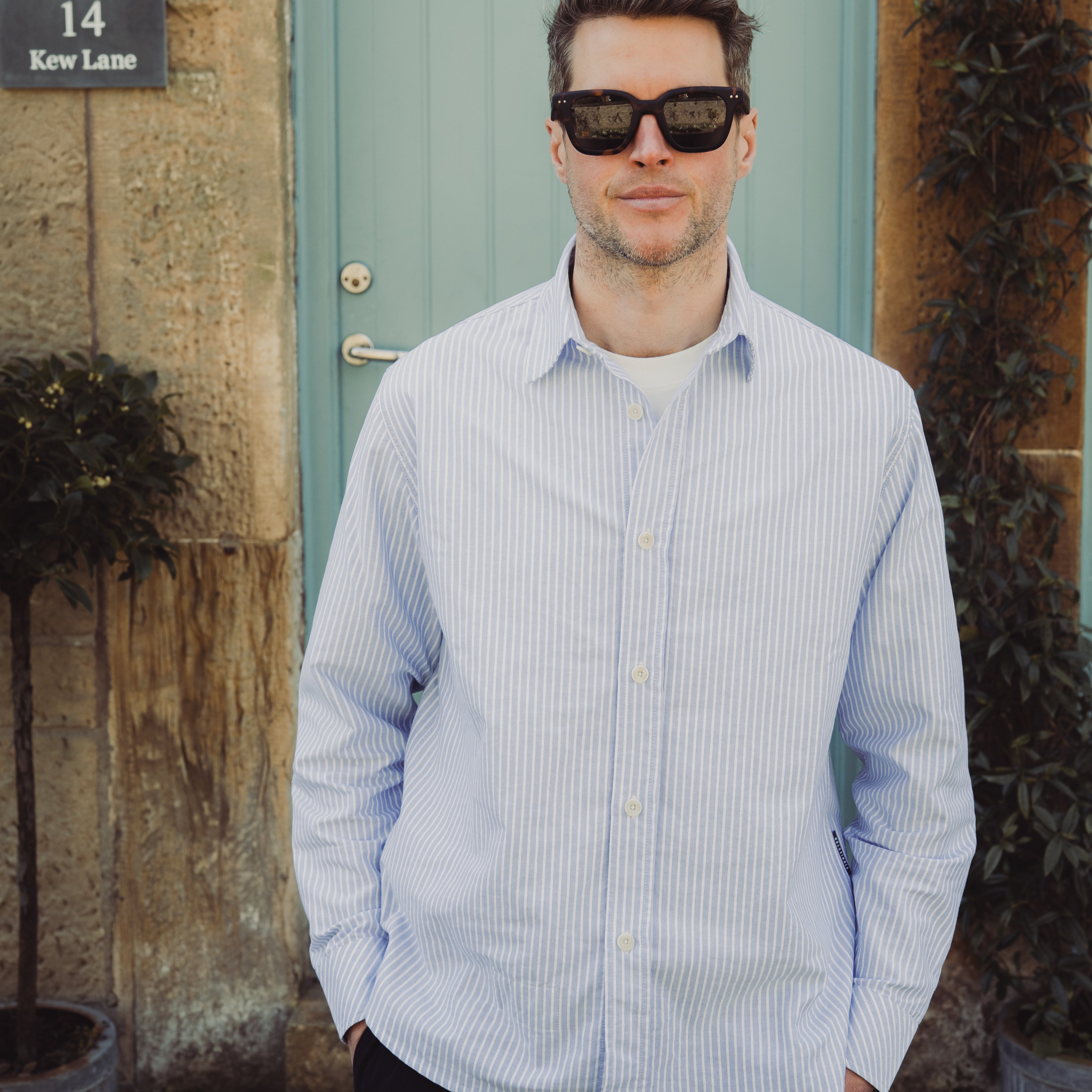 Man wearing sunglasses and a light blue striped shirt standing in front of a building with a door and plants.