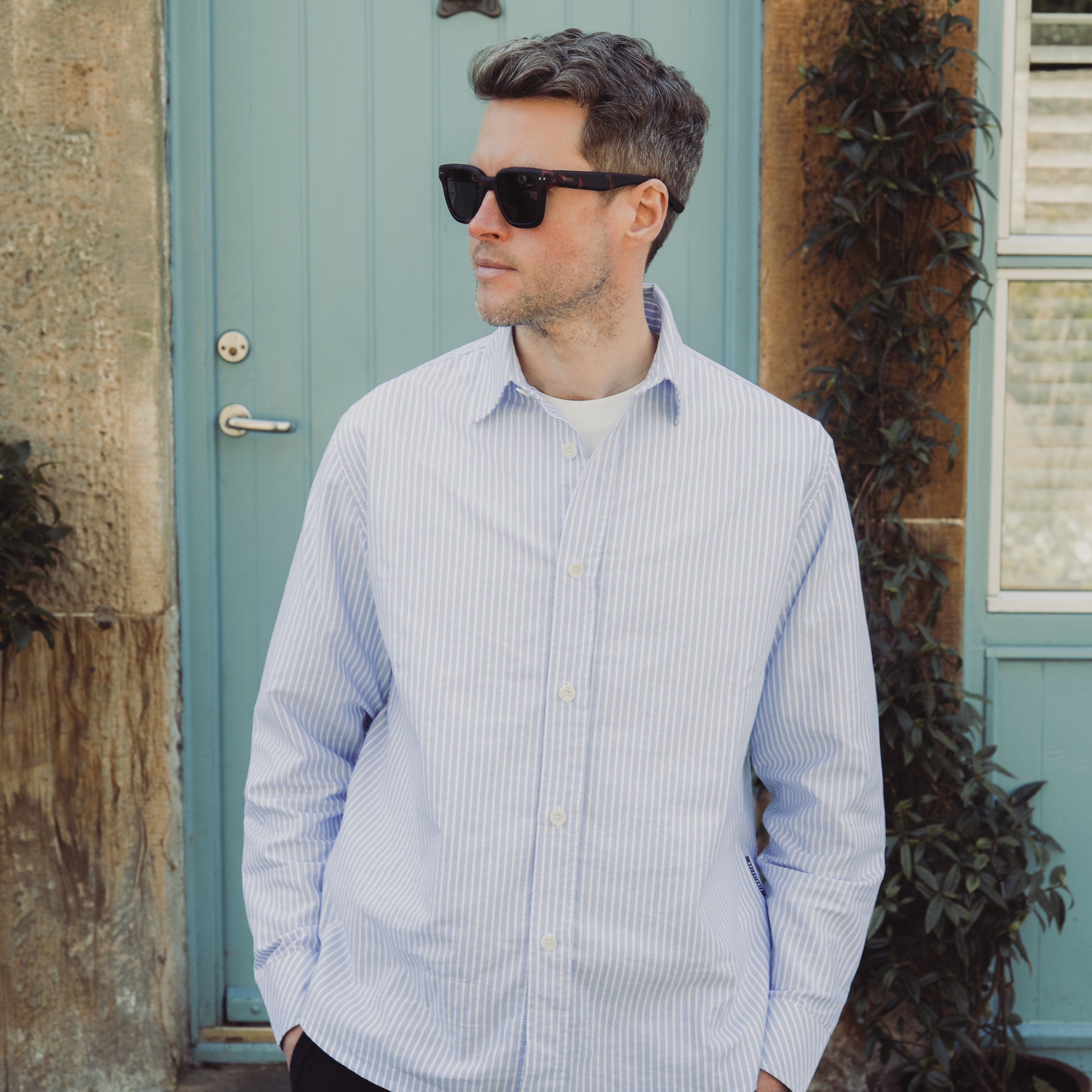 Man wearing a light blue striped shirt standing in front of a teal door.