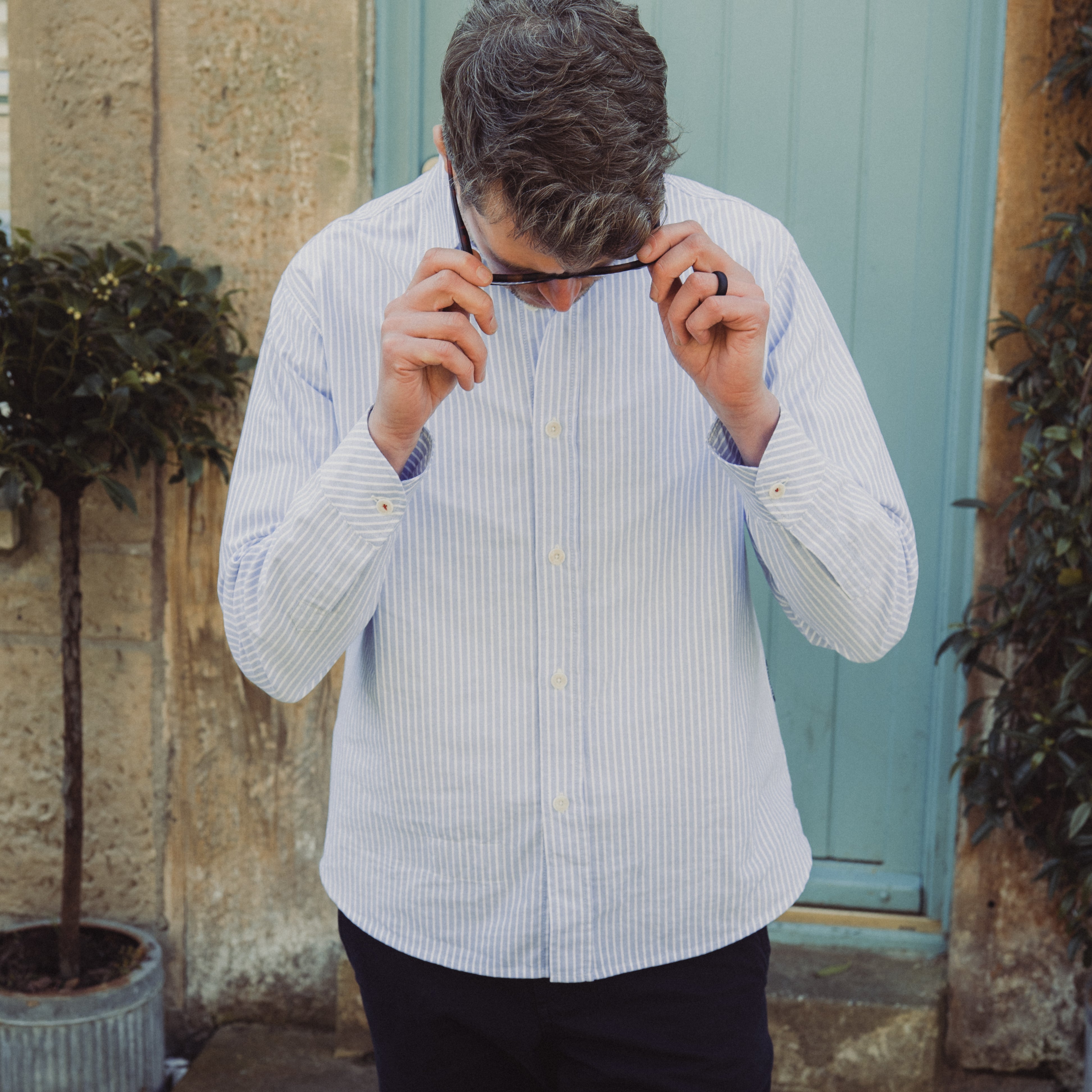 Man adjusting glasses outdoors against a stone wall and green door.