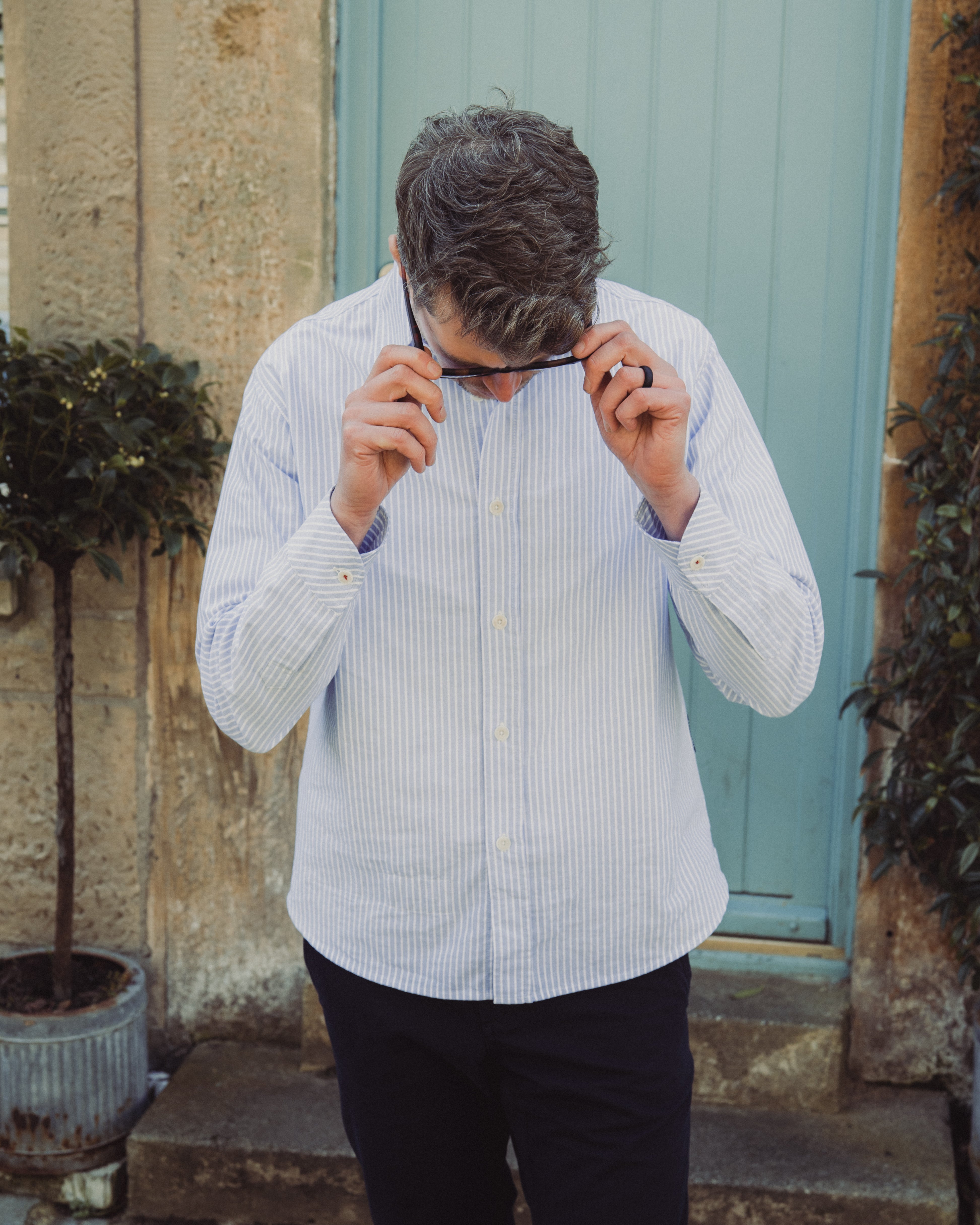 Man adjusting glasses outdoors against a stone wall and green door.