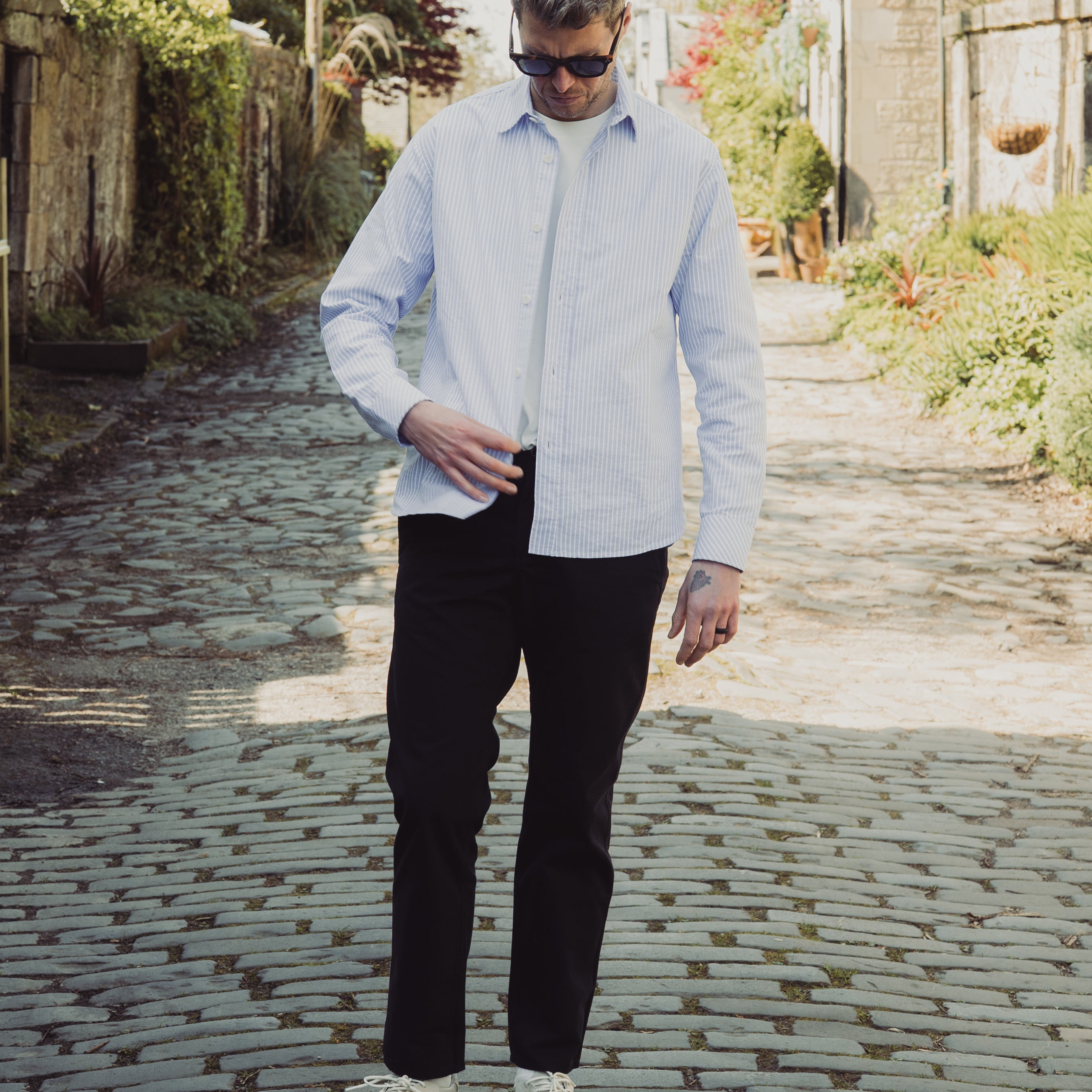 Man in light blue shirt and black pants standing on a cobblestone path with greenery around.